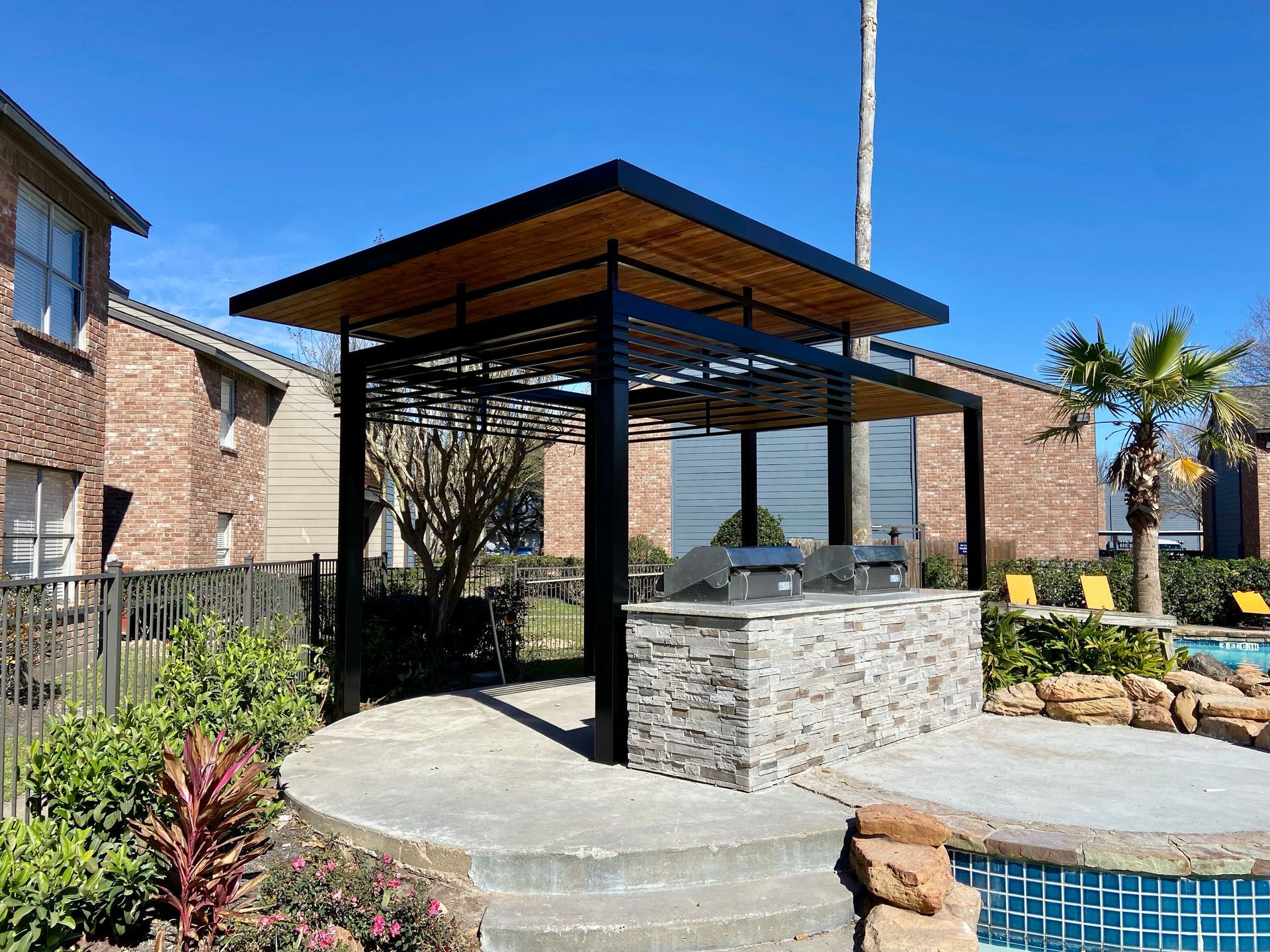Outdoor grilling area with a brick grill, black frame, and a wood and metal roof, near a pool.