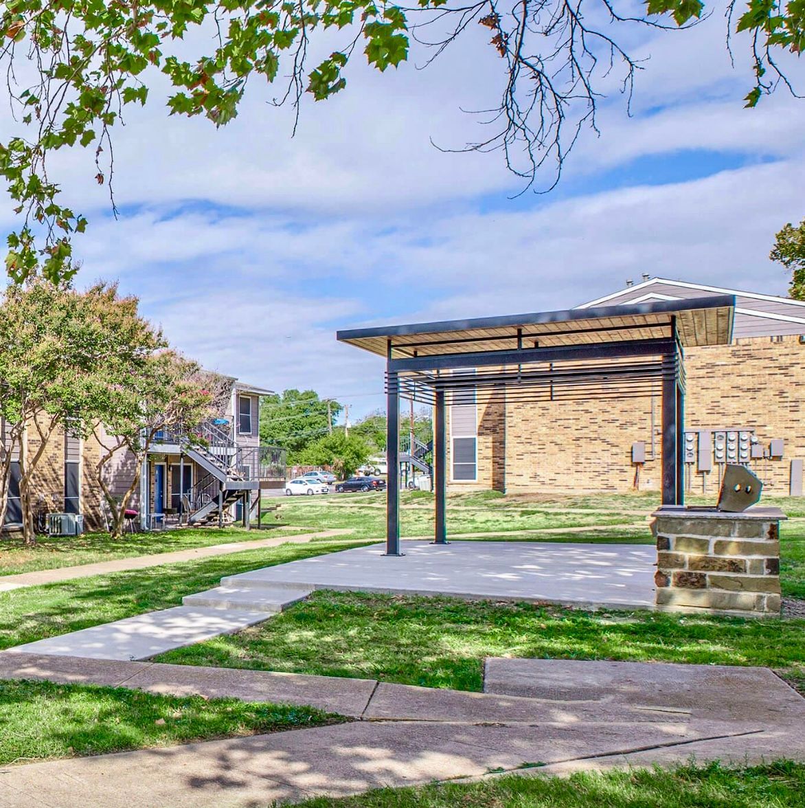 Park with shelter and brick structure; trees and buildings in background, bright day.
