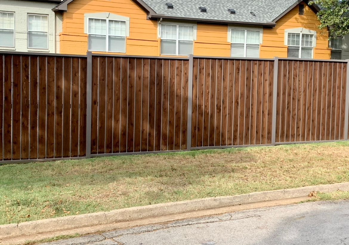 Brown wooden fence in front of a yellow brick house with white-framed windows on a grassy lawn.