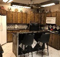A kitchen with wooden cabinets , stools , a refrigerator , microwave , and a ceiling fan.