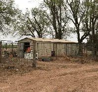 A shed in the middle of a dirt field with trees in the background.
