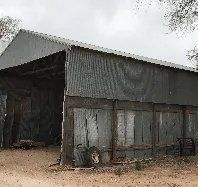 An old barn with a corrugated metal roof is sitting in the middle of a dirt field.