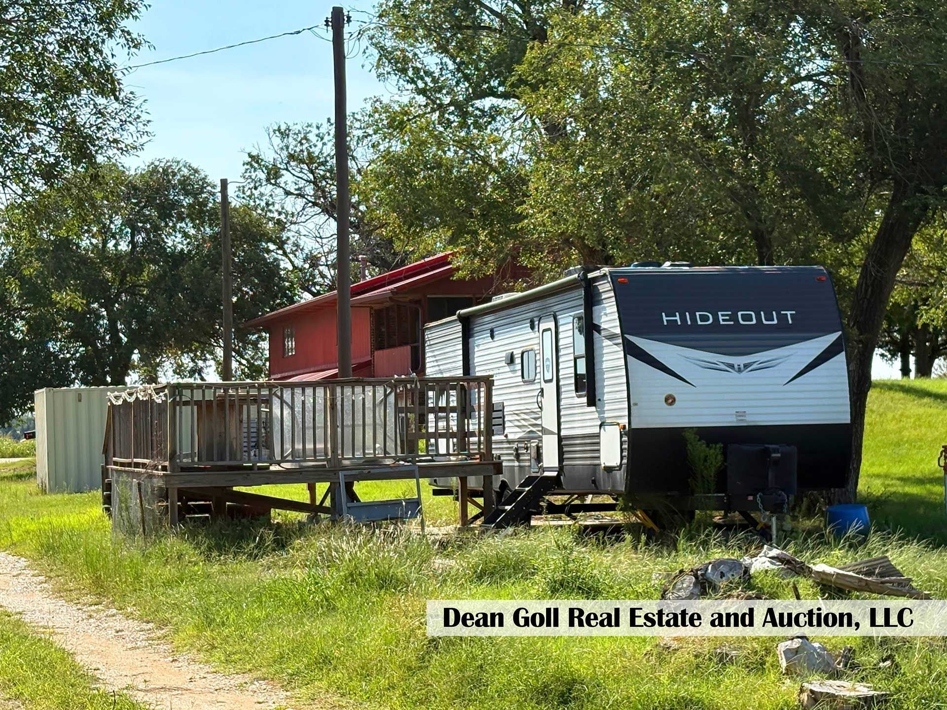 RV with deck, red building, trees, and grass in a rural outdoor setting
