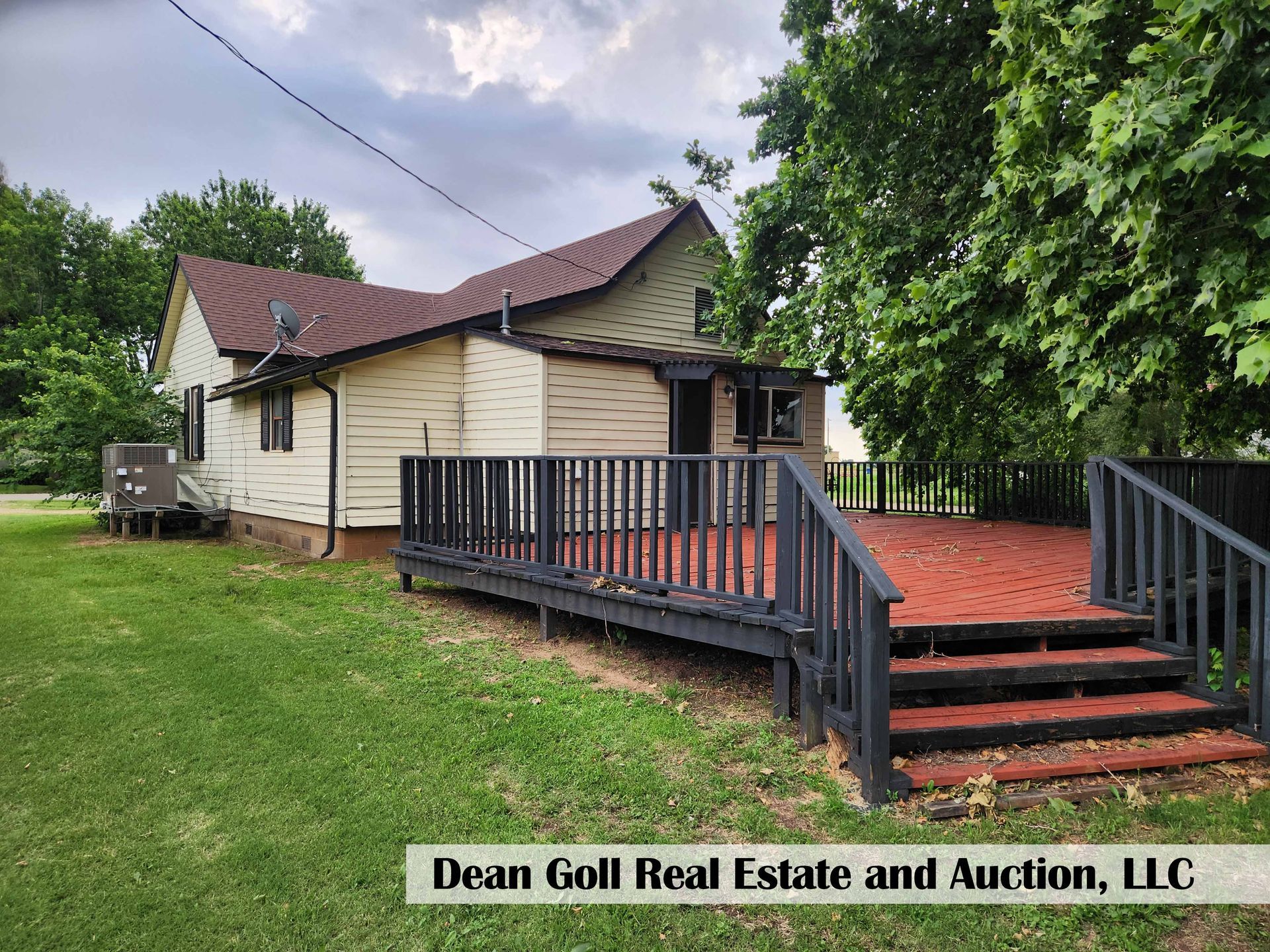 A two-story house with a large deck, surrounded by green grass and trees. The house is painted beige, and the deck is stained red with gray railings.