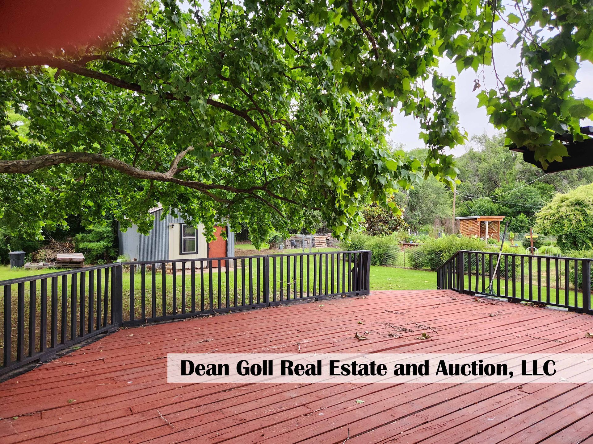 Red wooden deck with black railing overlooking a backyard with a shed and garden under a large tree.