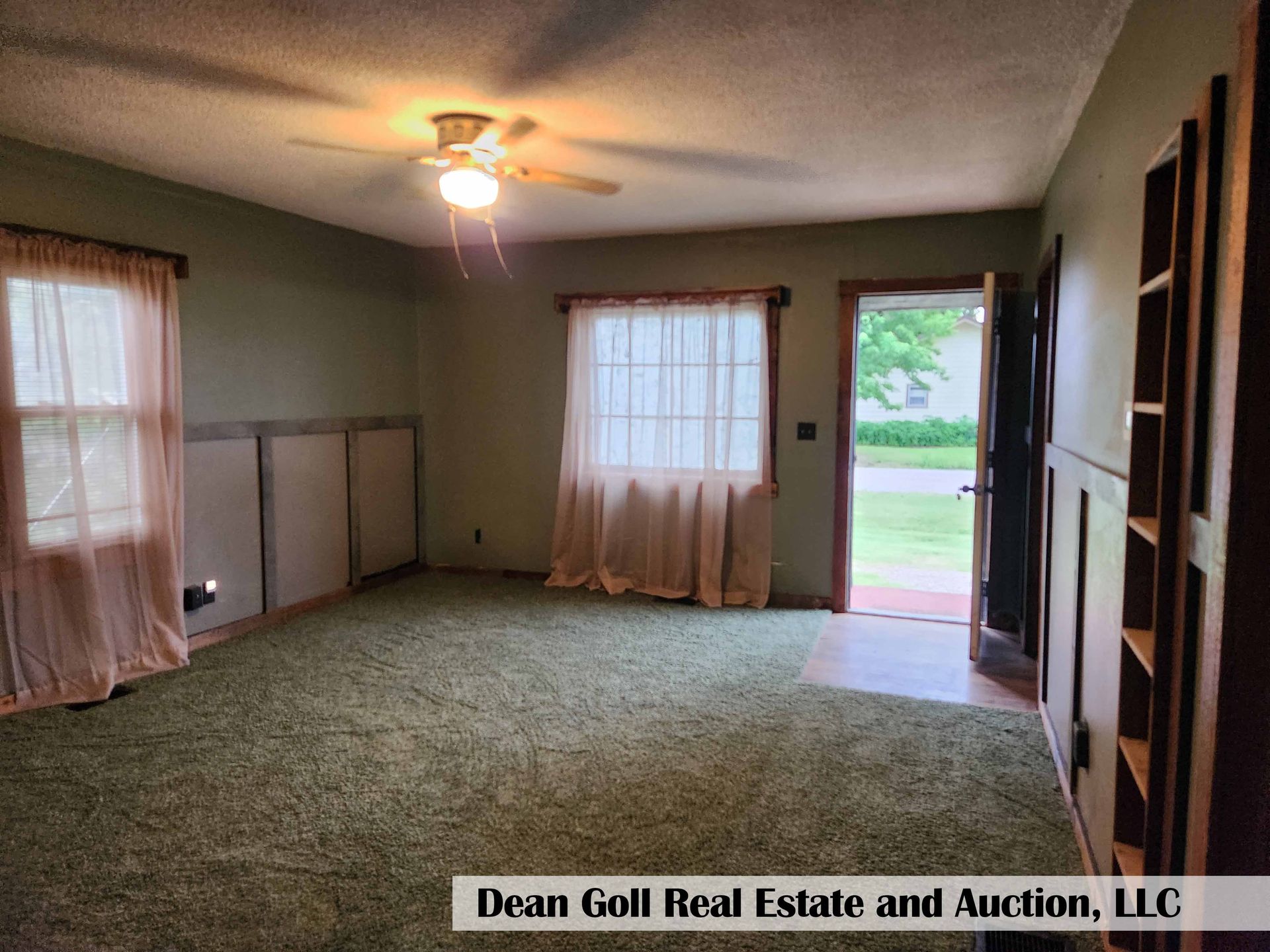 Living room with green carpet, sage green walls, peach curtains, and a built-in bookshelf. The door is open to the outdoors.