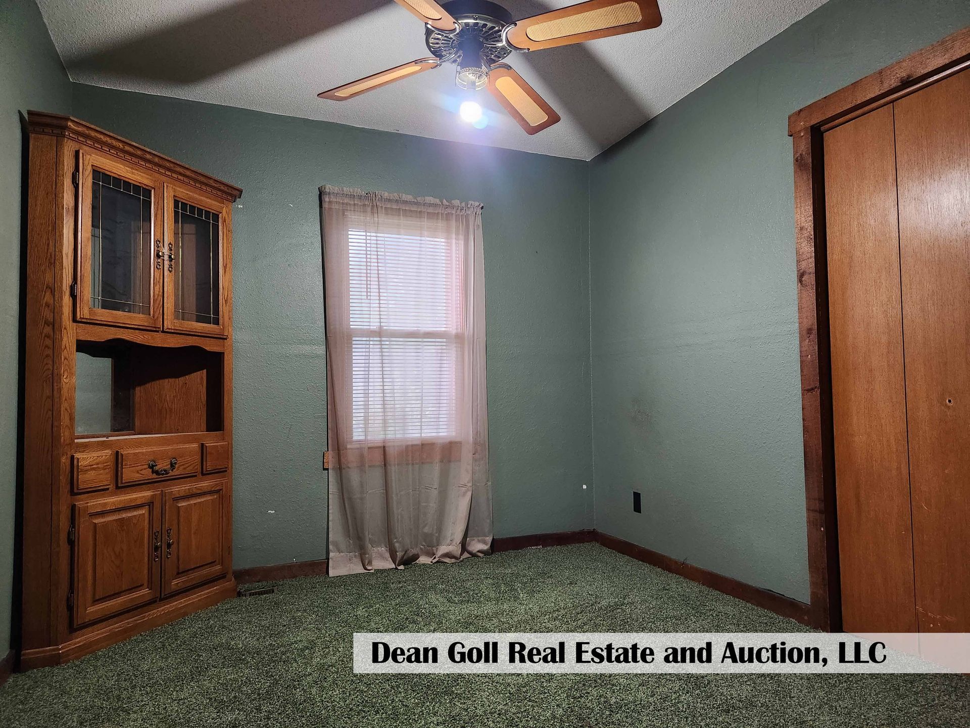 A small, empty room with green walls and a green carpet. A wooden cabinet and closed door are on opposite walls. A window with a sheer curtain is also present.