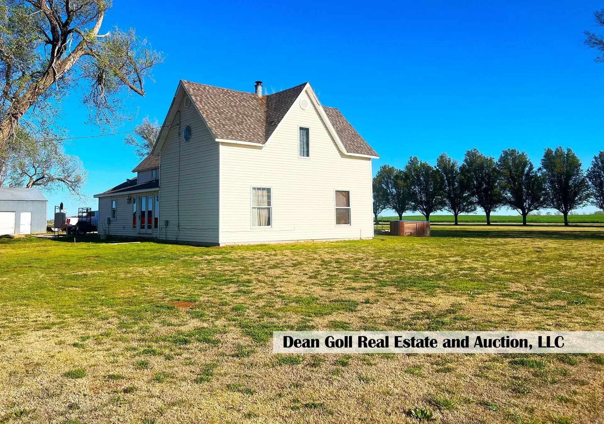 Old white farmhouse on a grassy field with a clear blue sky and a row of trees in the background.