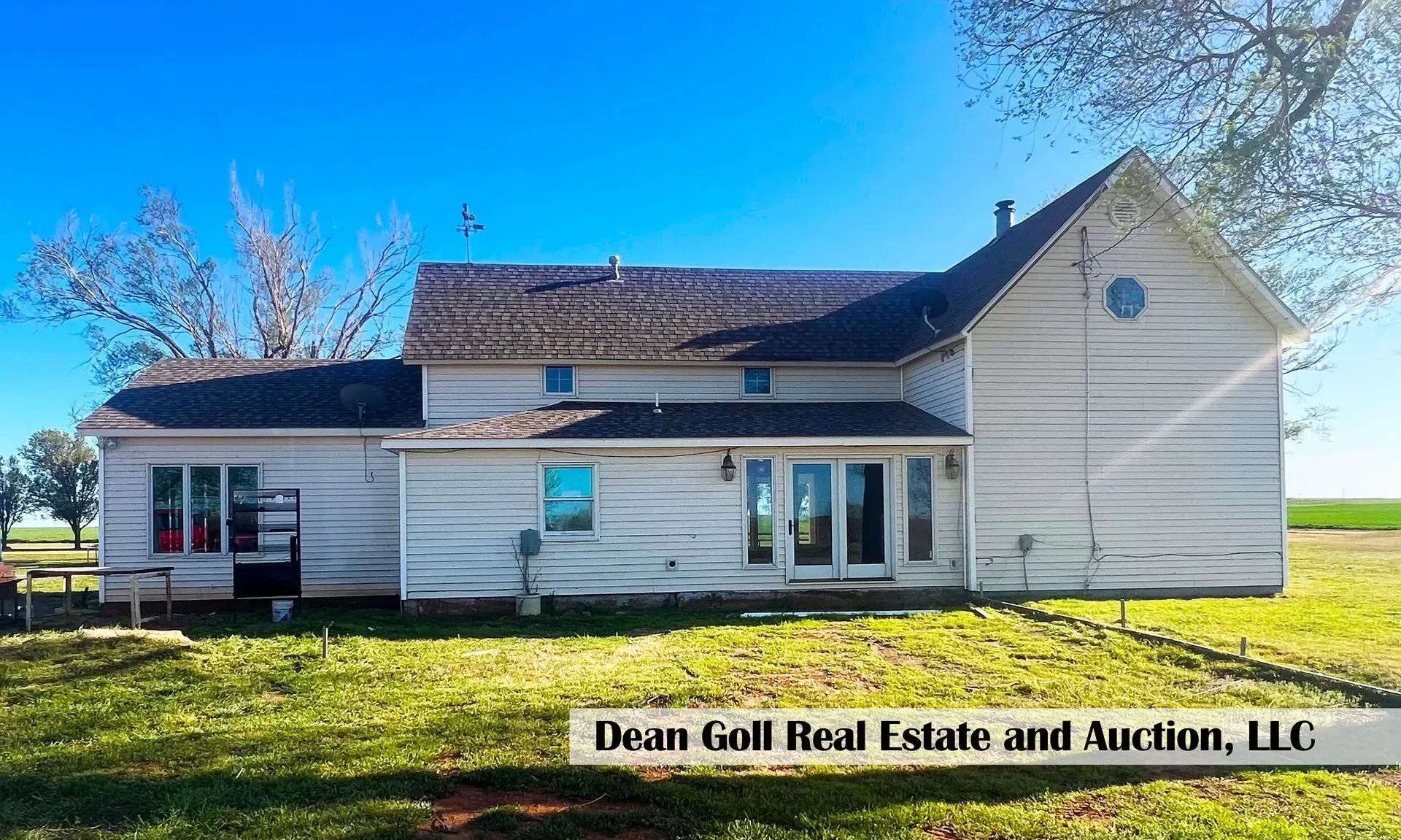 White farmhouse with visible weathered siding and a dark roof, set in a grassy field under a clear blue sky.