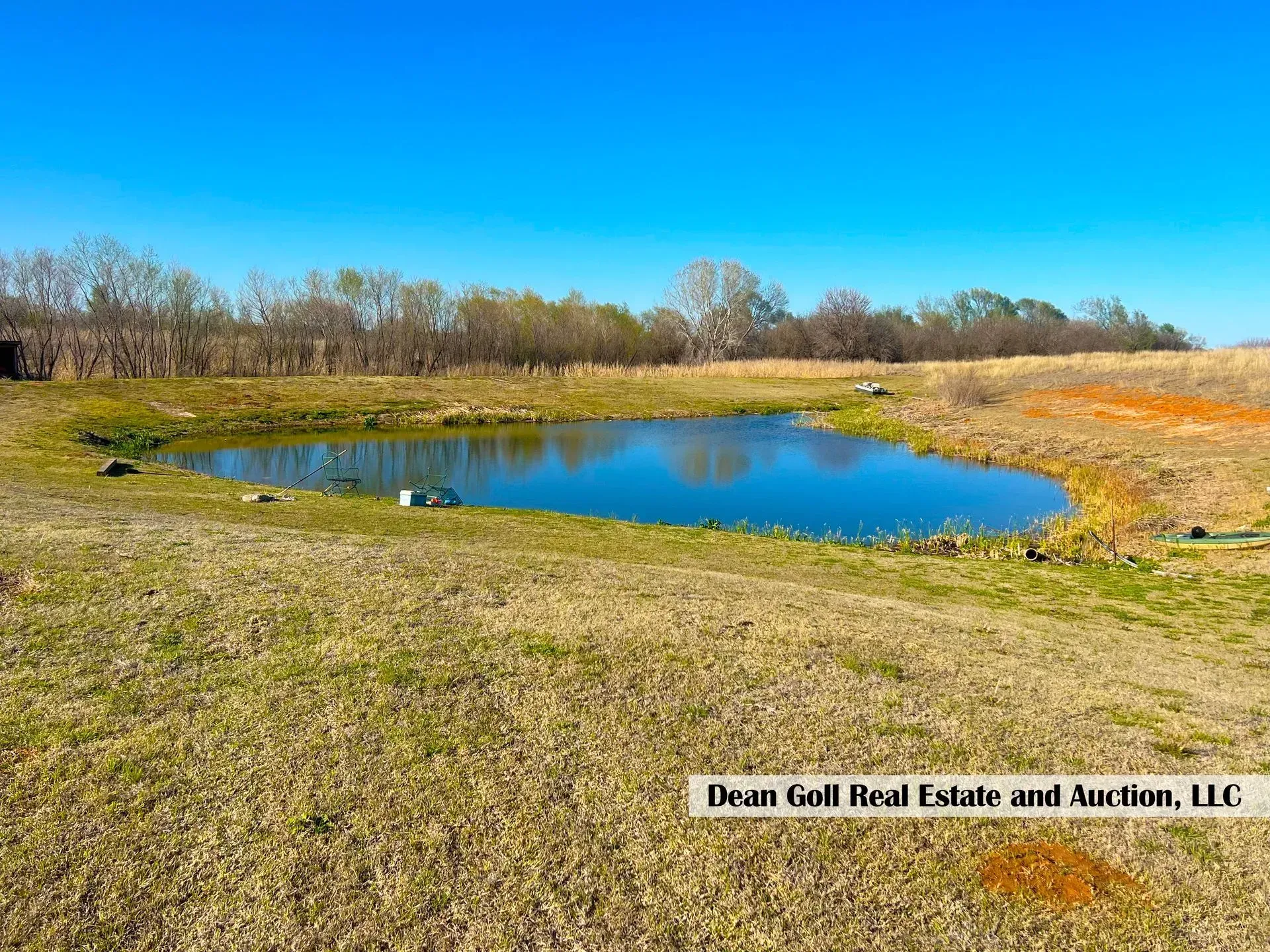 A small pond surrounded by dry grass and a line of trees under a clear blue sky.