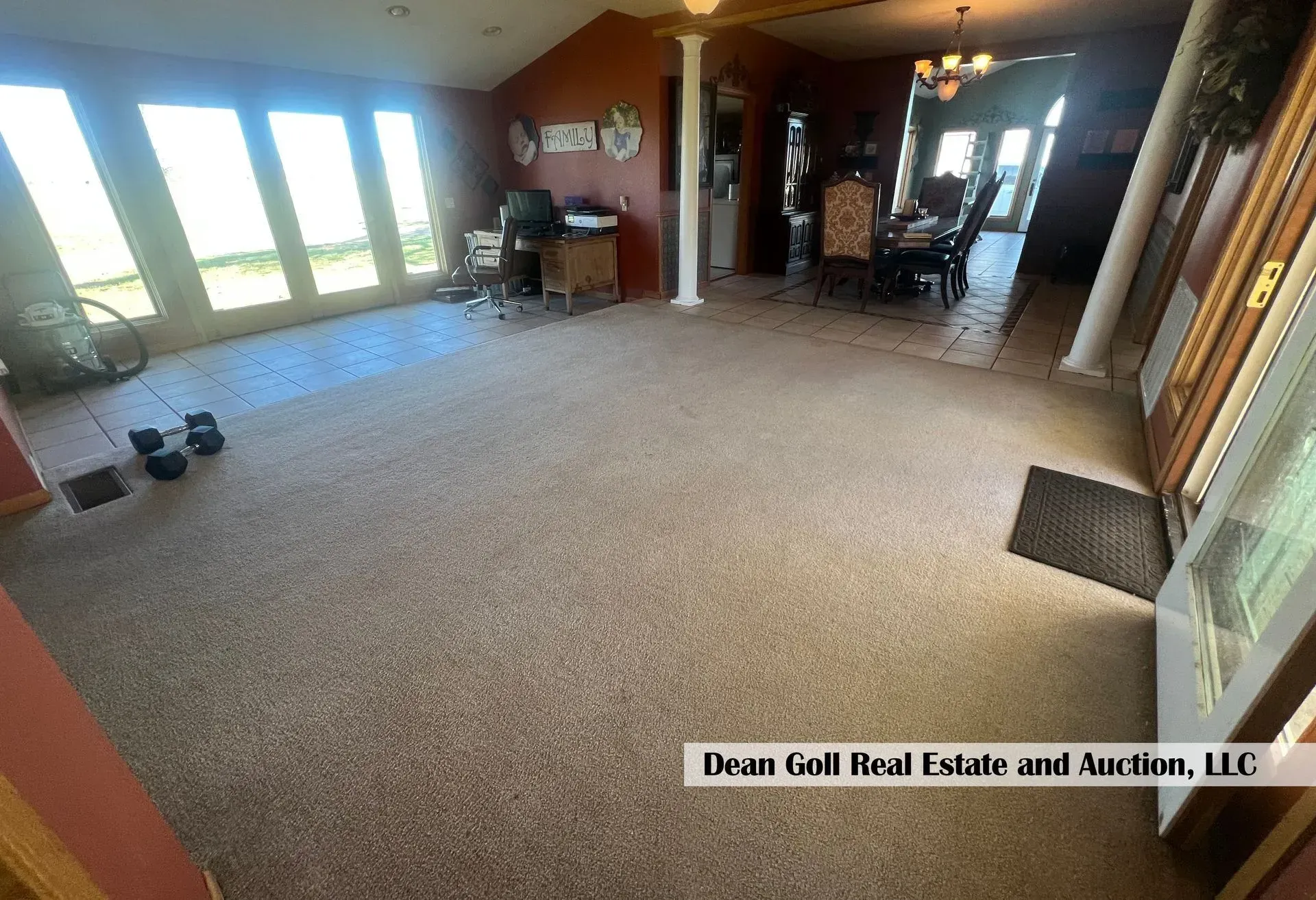 Spacious living area with large windows, columns, and a dining area in the background. Brown carpet and walls.