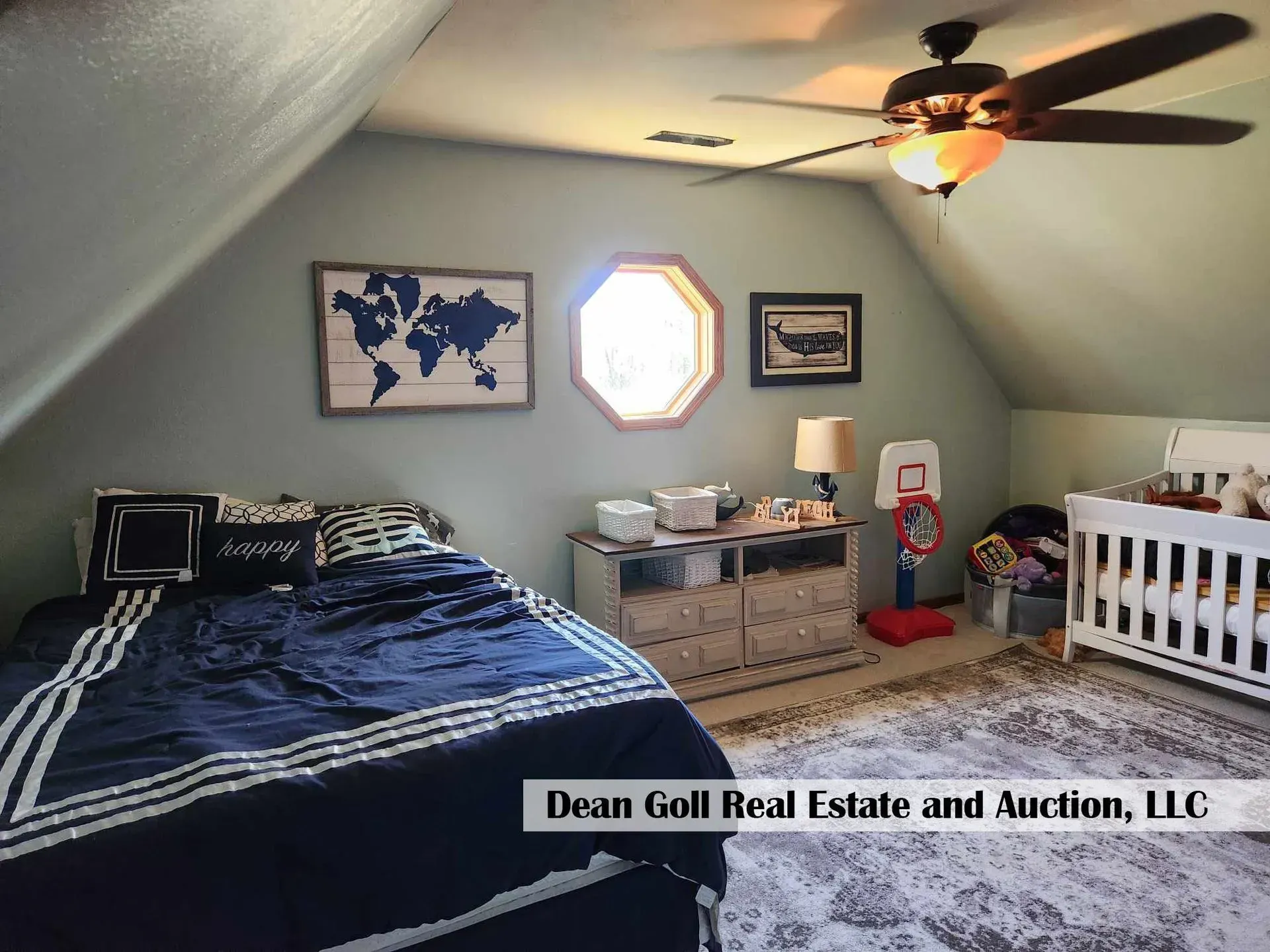 Bedroom with slanted ceilings, bed, dresser, crib, and a ceiling fan. The walls are light blue with wooden artwork.