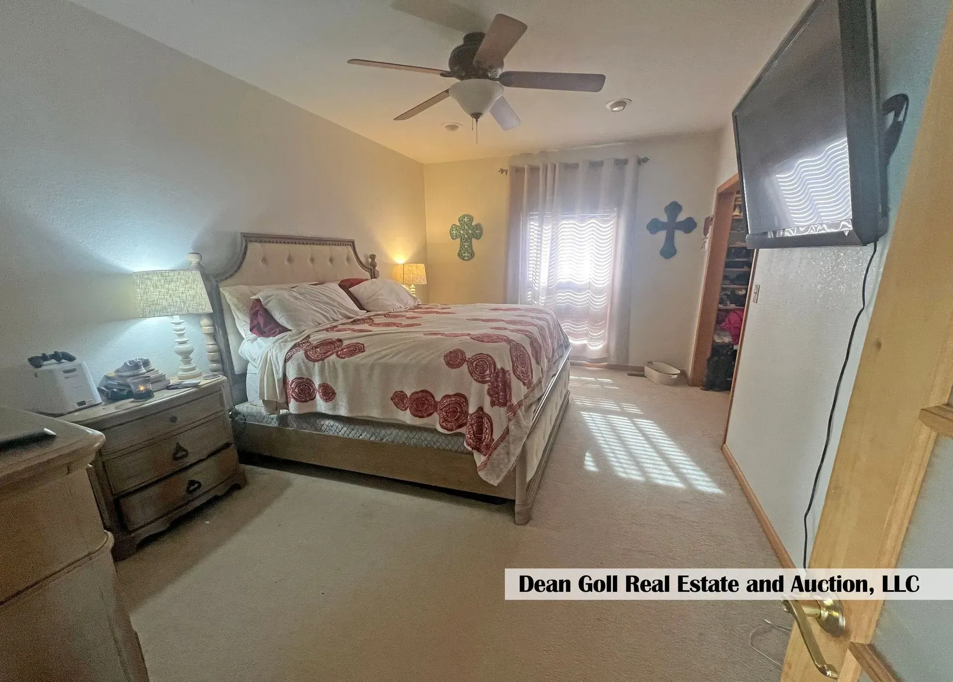 Bedroom with a bed, nightstand, TV, ceiling fan, and a doorway to a balcony. Light-colored walls and carpet.