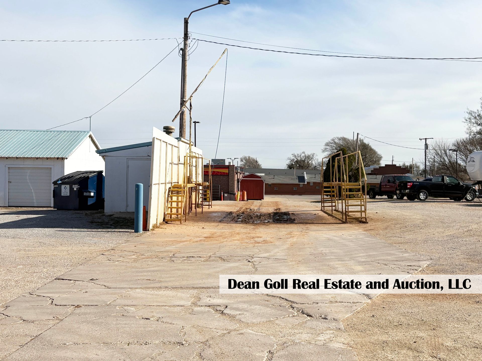 A gravel lot with a scale, small buildings, power lines, and a truck, under a cloudy sky.