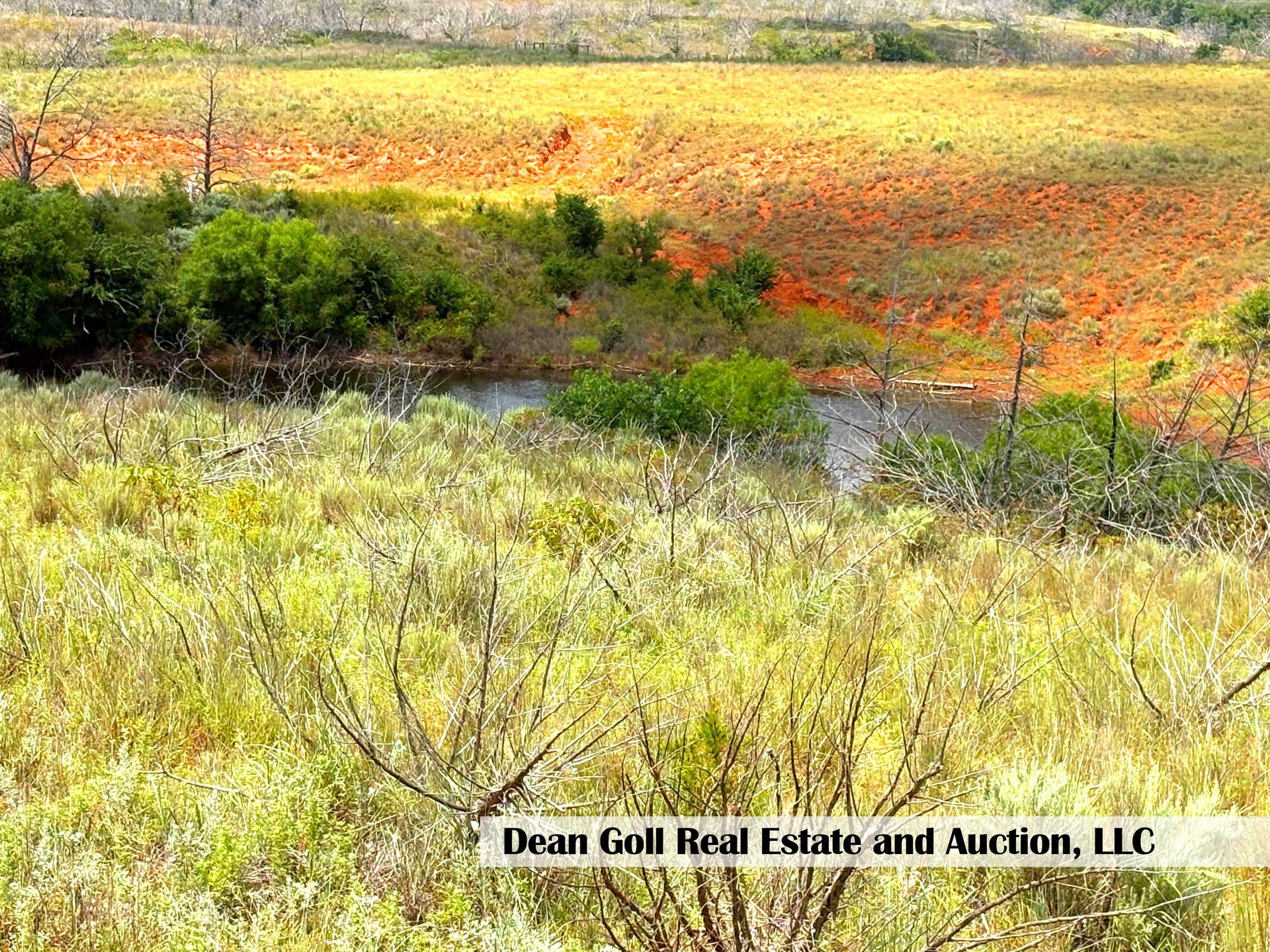 A landscape with a grassy foreground, a small body of water, and a reddish-brown field in the background under a sunny sky.