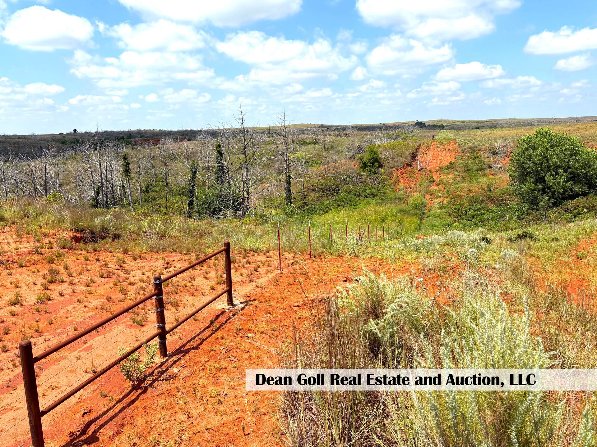 Red dirt and grassy plains with a fence in the foreground, under a blue sky with scattered clouds.