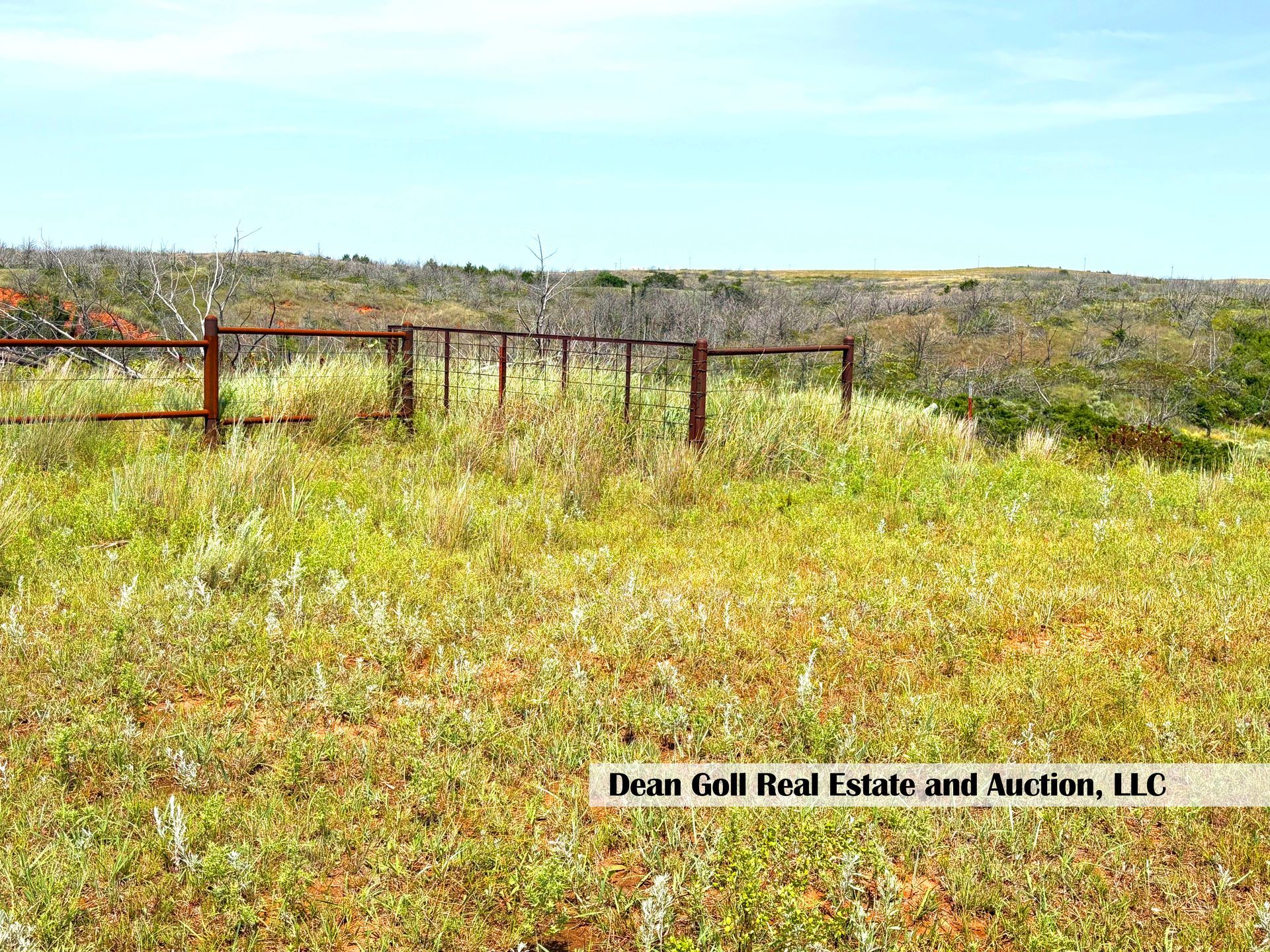 A rusty metal fence in a grassy field under a blue sky. The landscape appears to be a rural, undeveloped area.