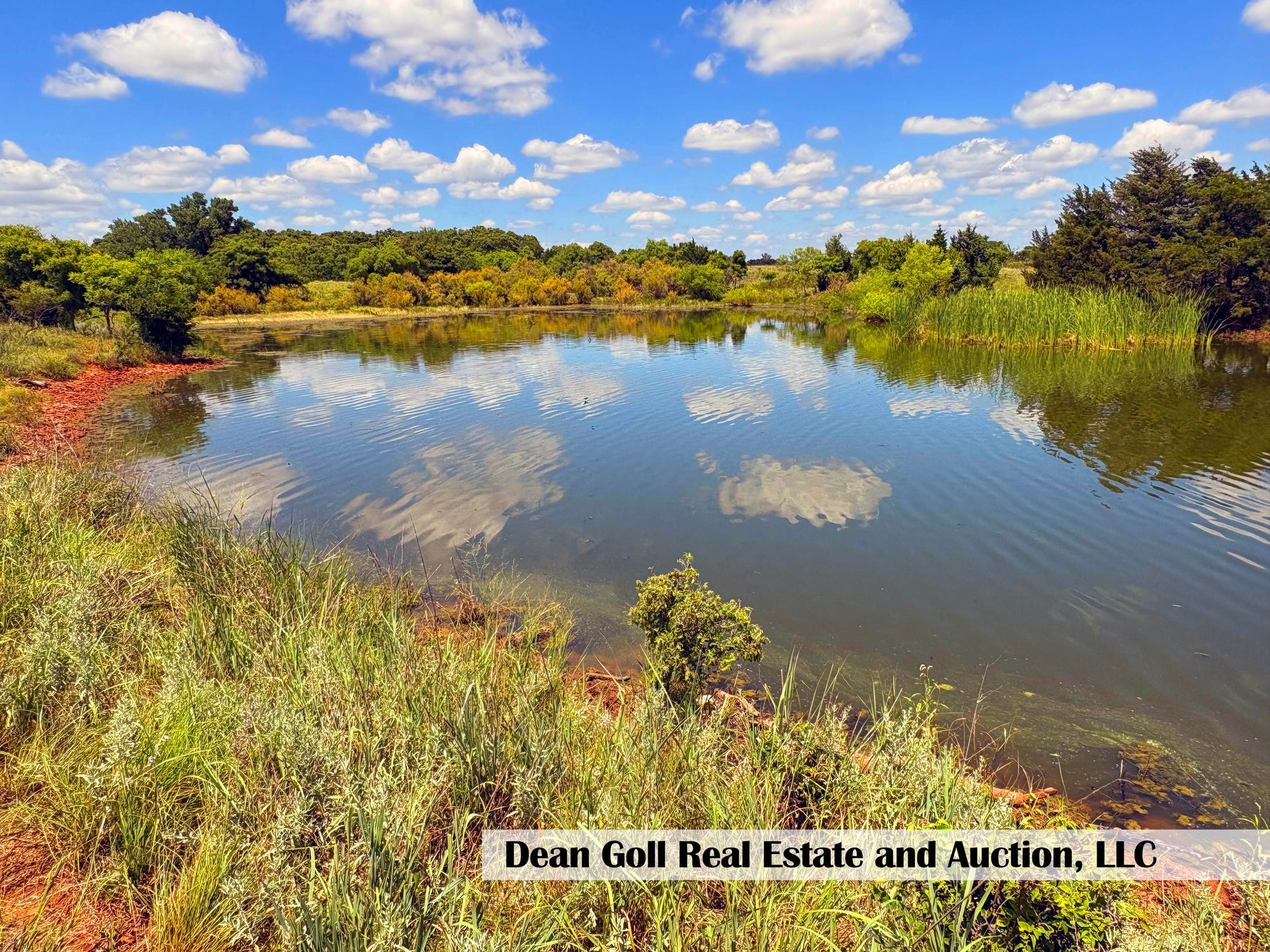 A calm lake reflects puffy clouds and blue sky. Lush vegetation lines the red-soiled banks with trees in the background.