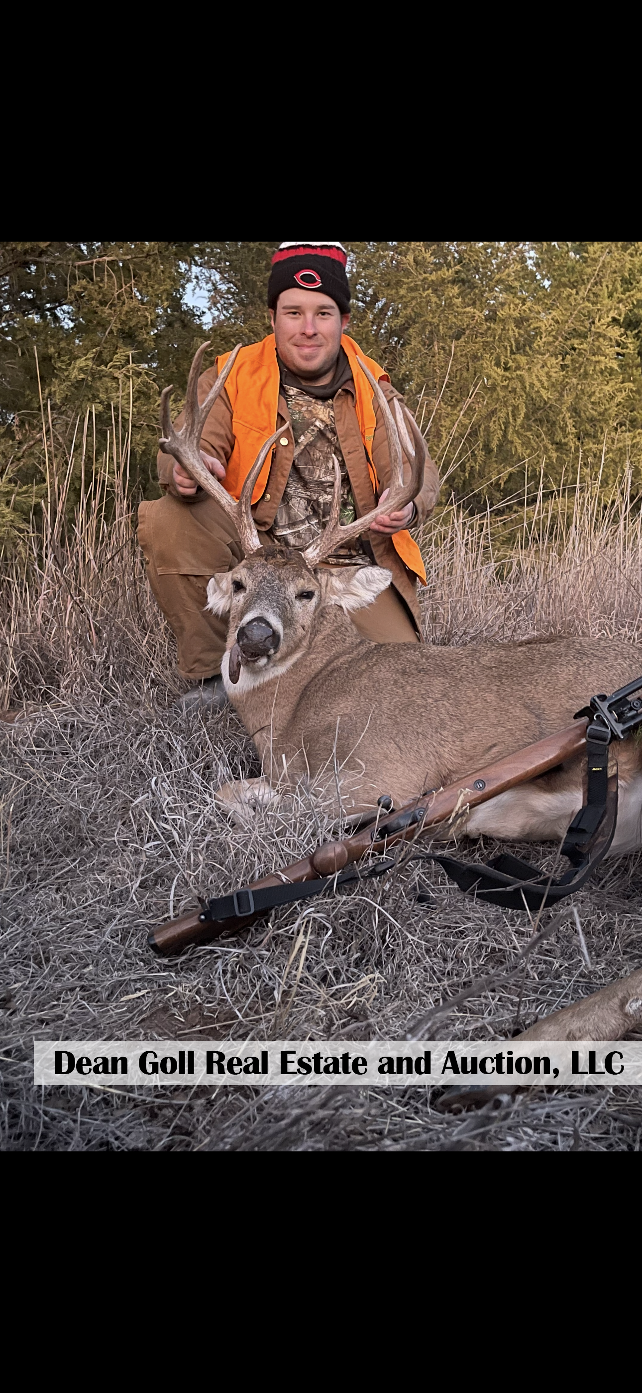 A man in camouflage and orange vest kneels next to a large deer he has shot. The deer is lying on the ground near a rifle.