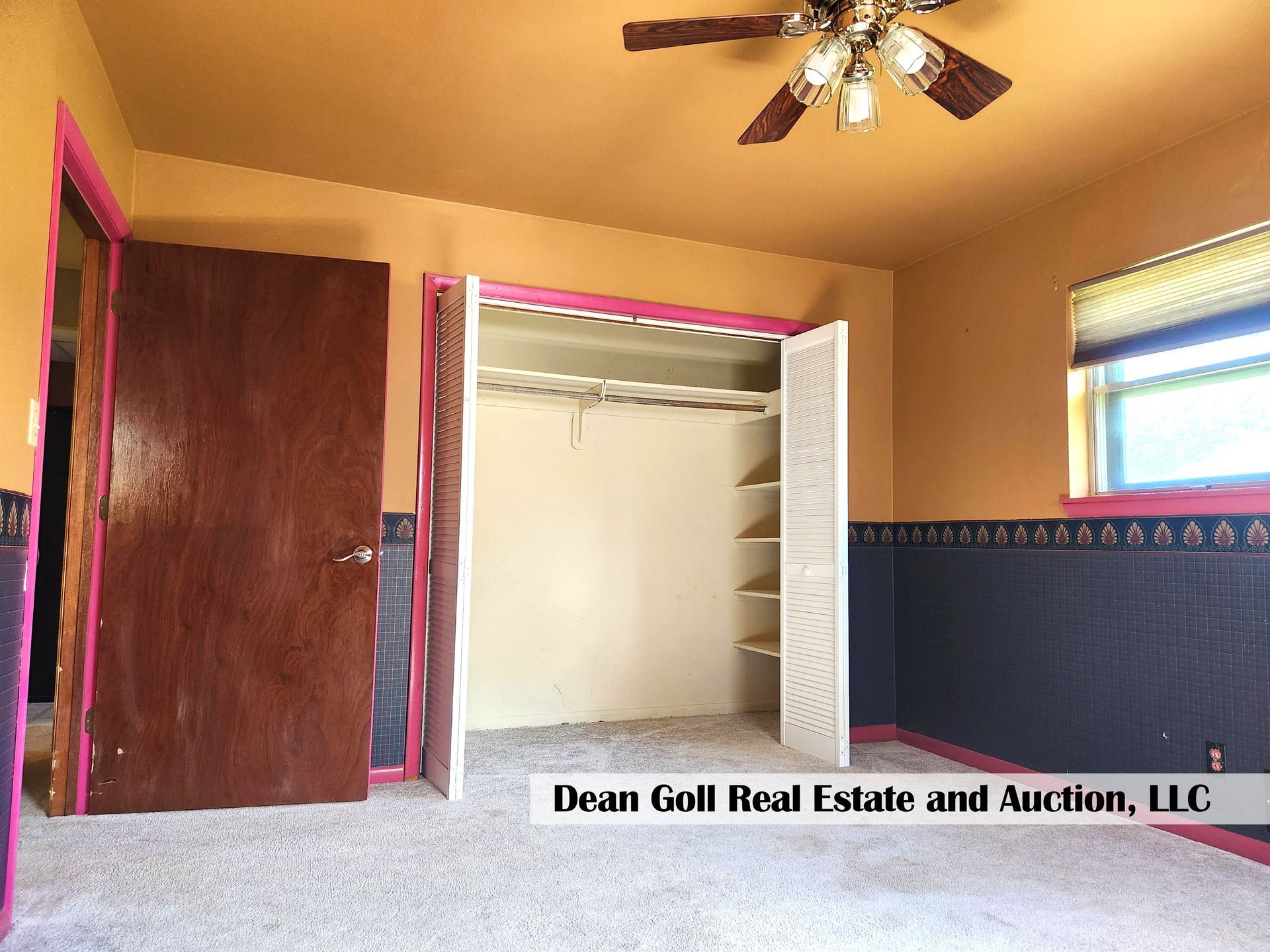 Empty bedroom with tan walls, pink trim, and a bi-fold closet. A ceiling fan hangs above, and a window is on the right.