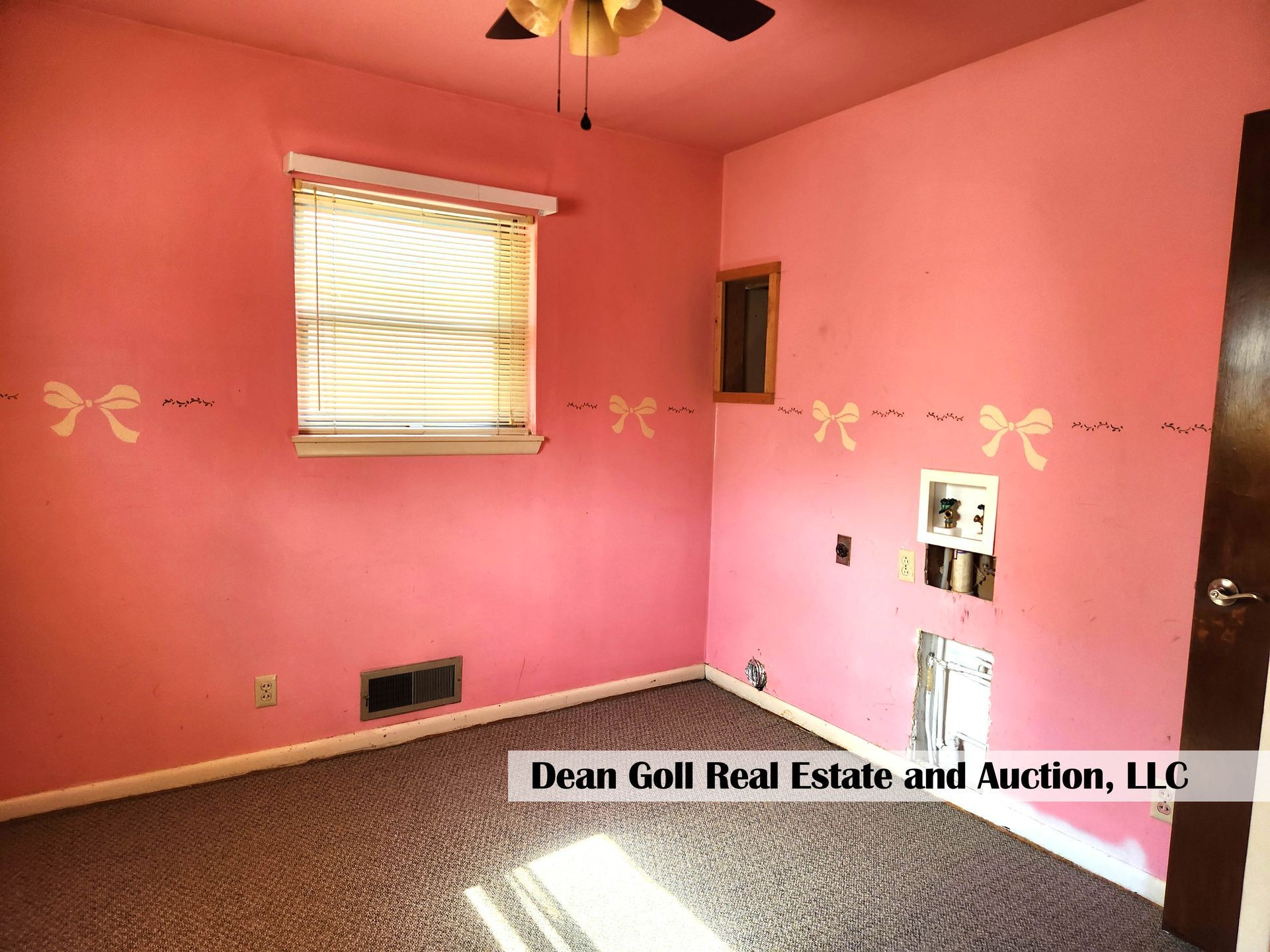 A pink-painted bedroom with a bow stencil design, carpet, and a window. There are exposed wires in the wall.