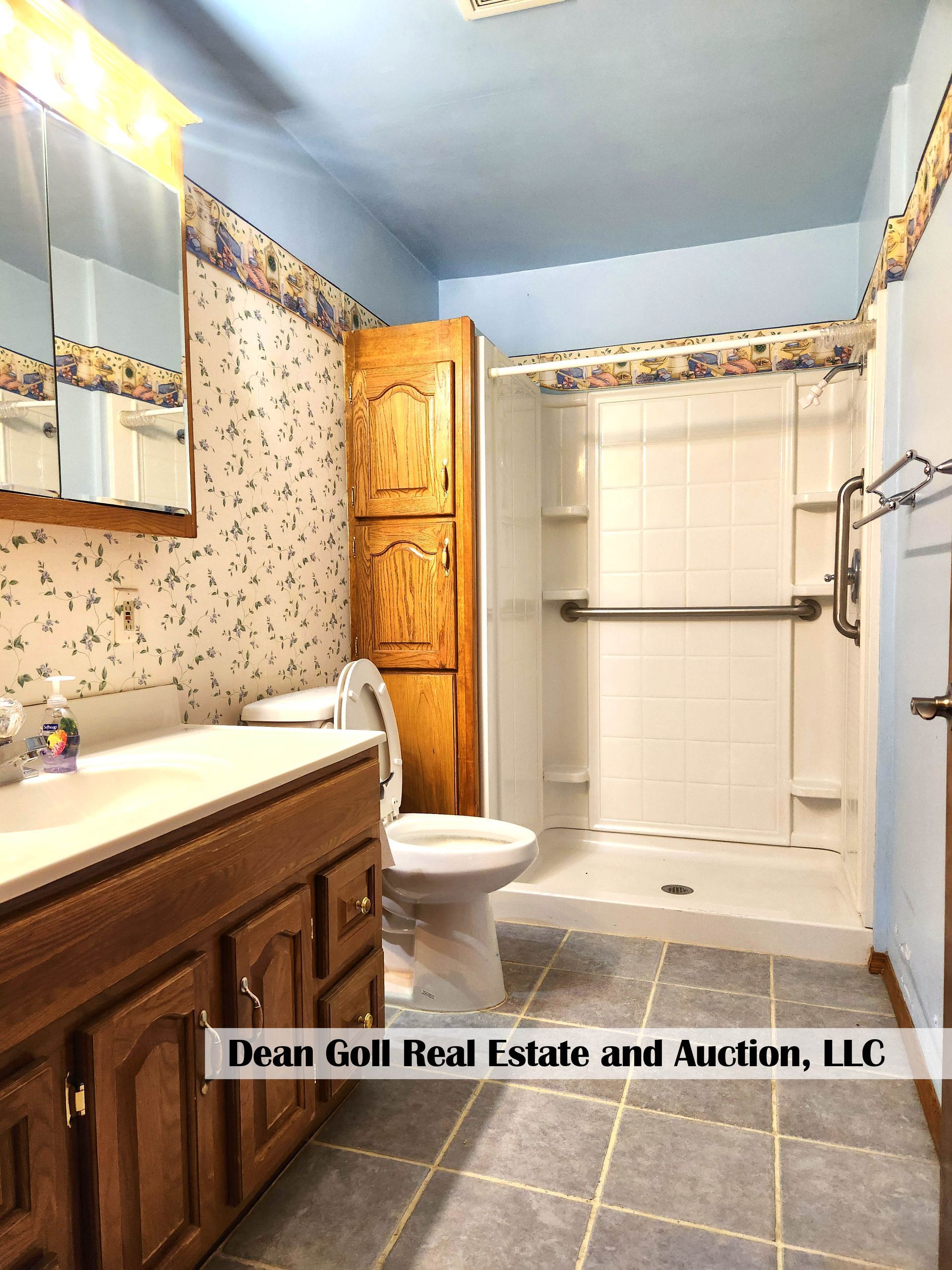 Bathroom with blue ceiling, patterned wallpaper, a wooden vanity, a toilet, and a shower stall with a grab bar.