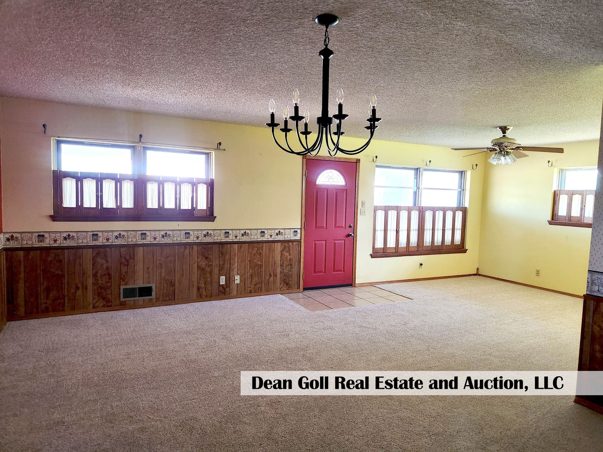 Interior of a room with beige carpet, wood paneling, yellow walls, and a red door. A chandelier hangs from the textured ceiling.