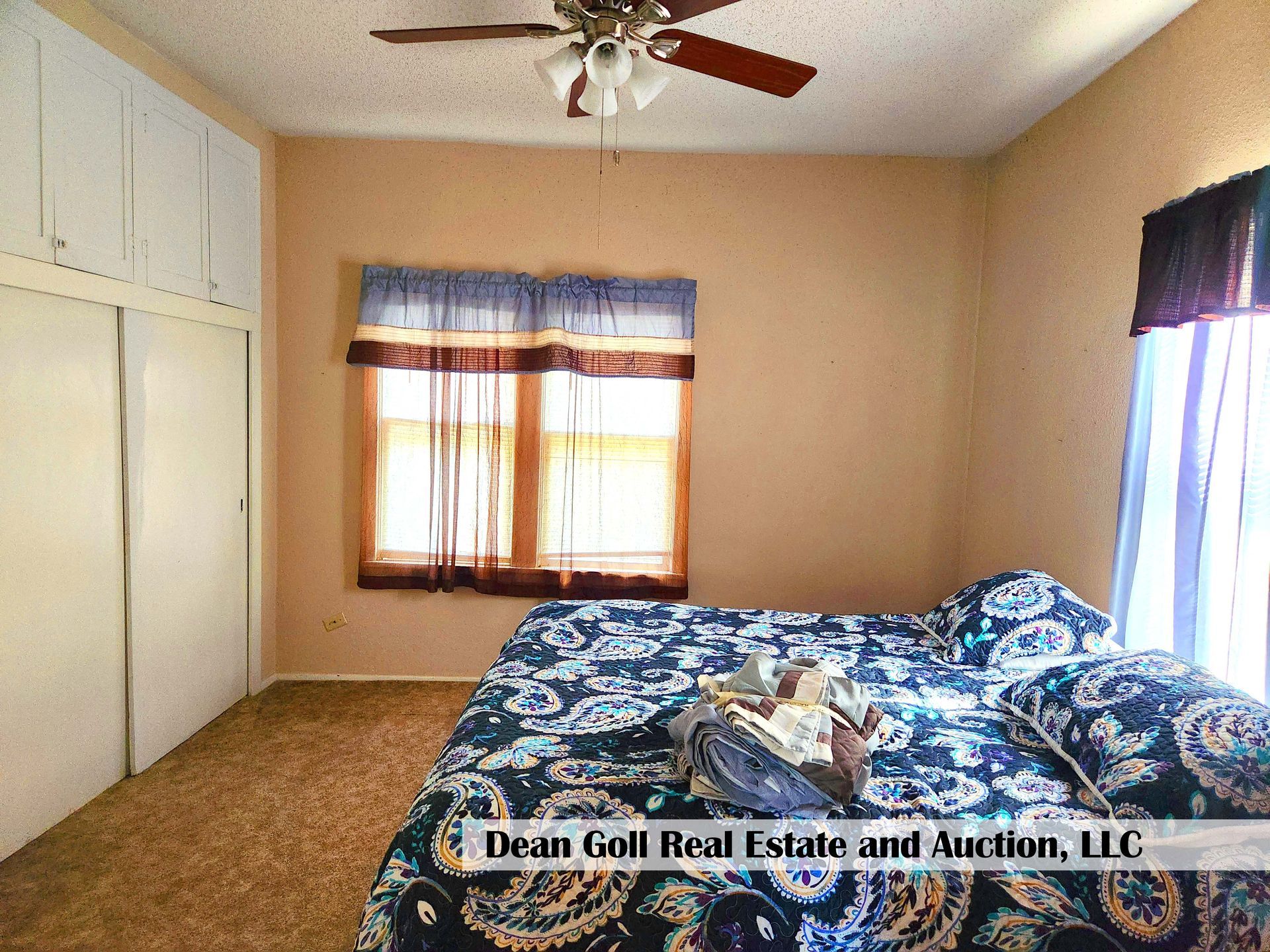 Bedroom with bed, window with patterned curtains, closet, and ceiling fan. Beige walls and carpet.