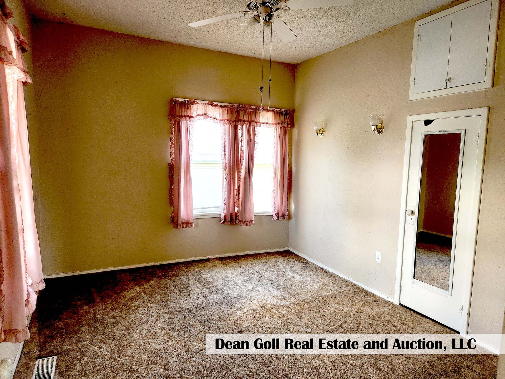 A room with brown carpet, tan walls, and two windows with pink curtains. A mirror is on the door.