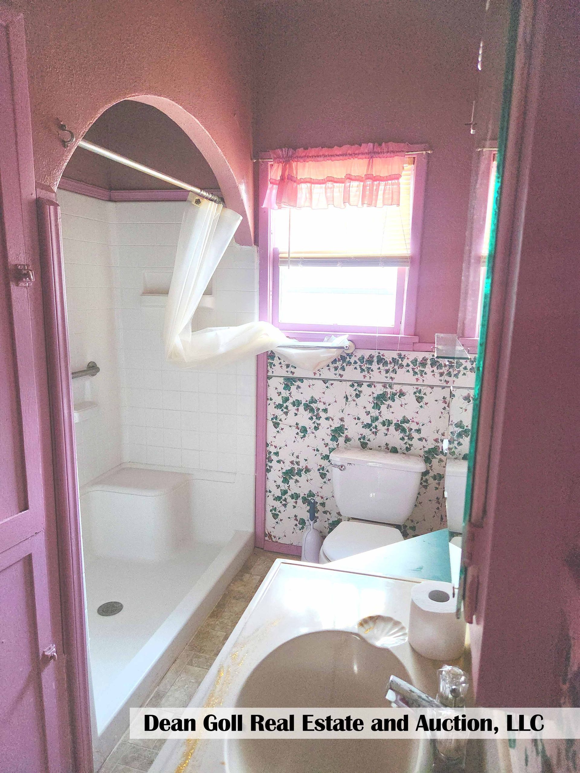 A small, pink bathroom with a shower, toilet, and sink. The walls have patterned wallpaper and a window with a pink curtain.