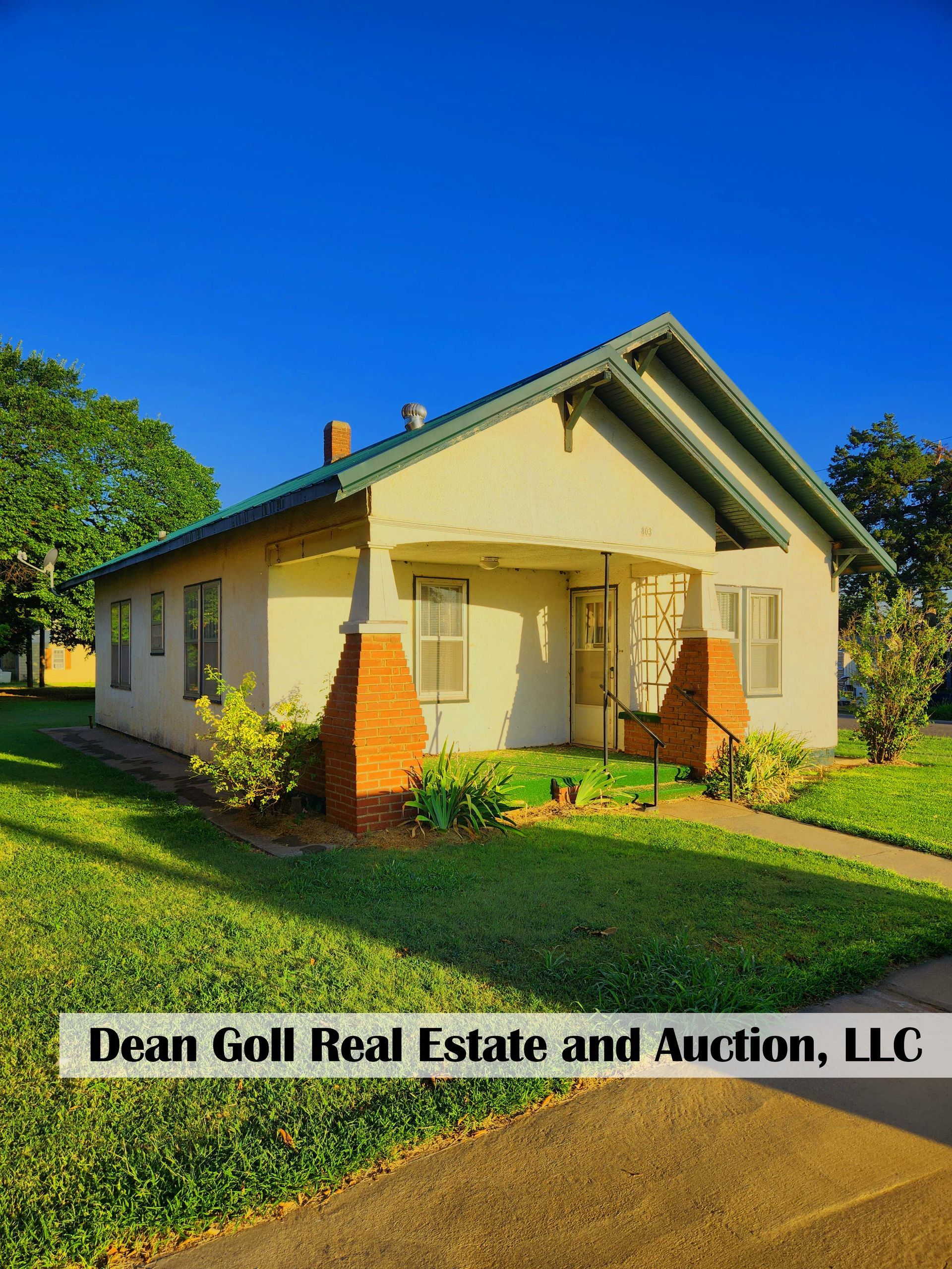 A light-colored bungalow with a green roof and red brick supports, set on a green lawn under a clear blue sky.