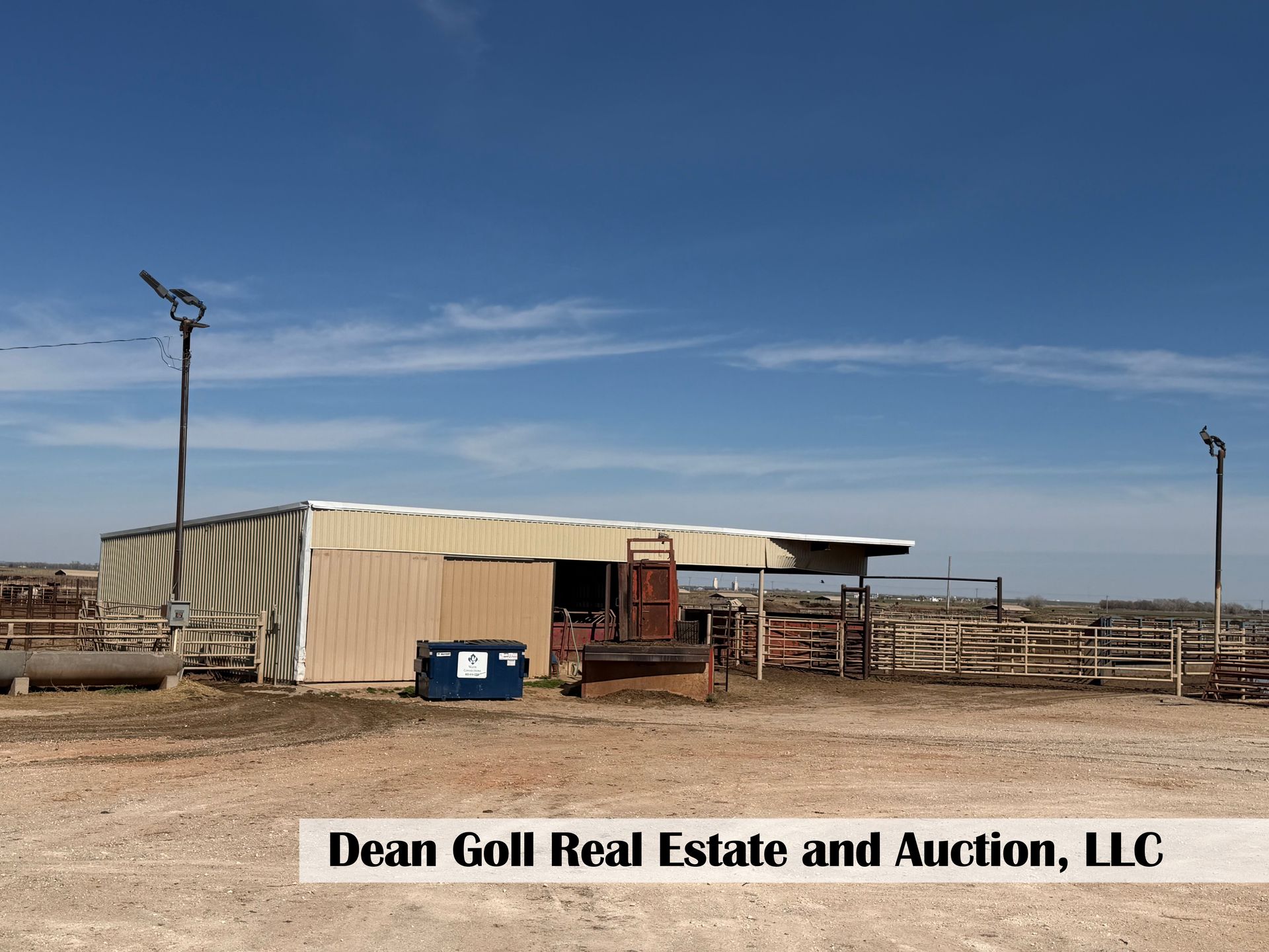 A tan metal barn with an open side and cattle pens on a dirt lot under a clear blue sky.