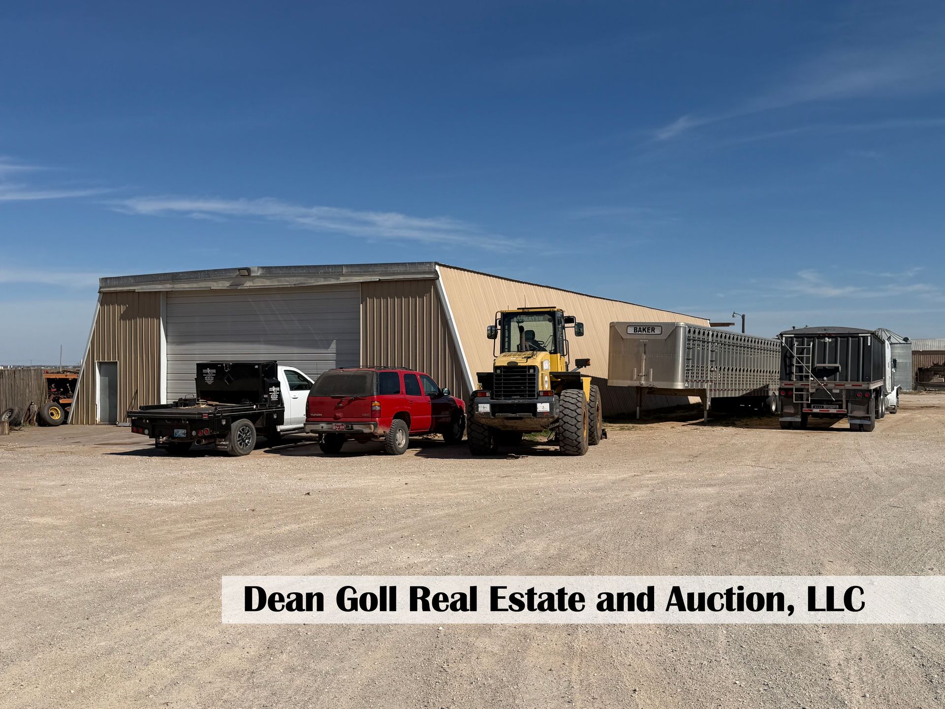 A tan warehouse with a construction vehicle and several trucks parked in a gravel lot under a clear blue sky.