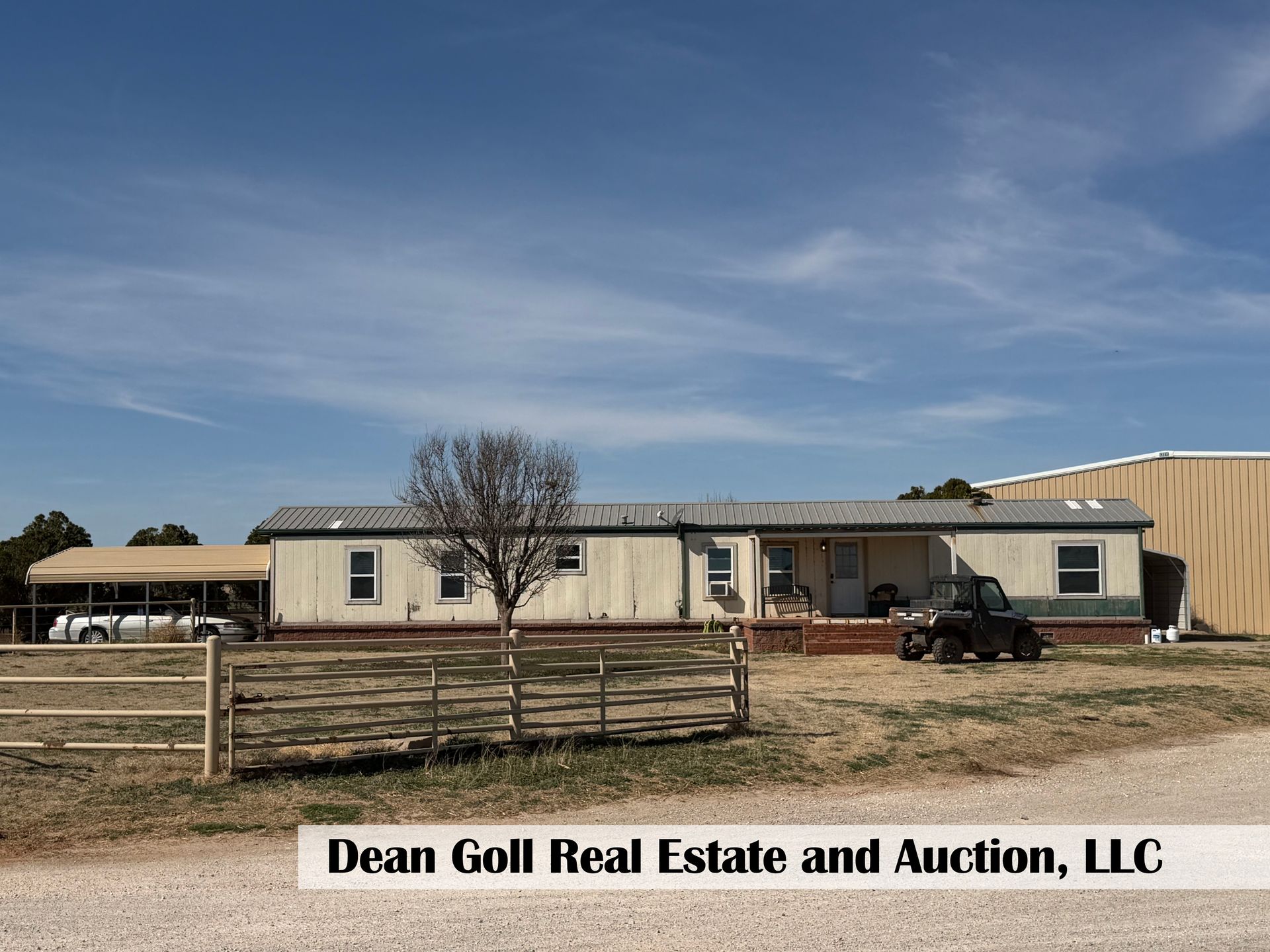 A long, light-colored mobile home with a carport and adjacent metal building under a blue sky.