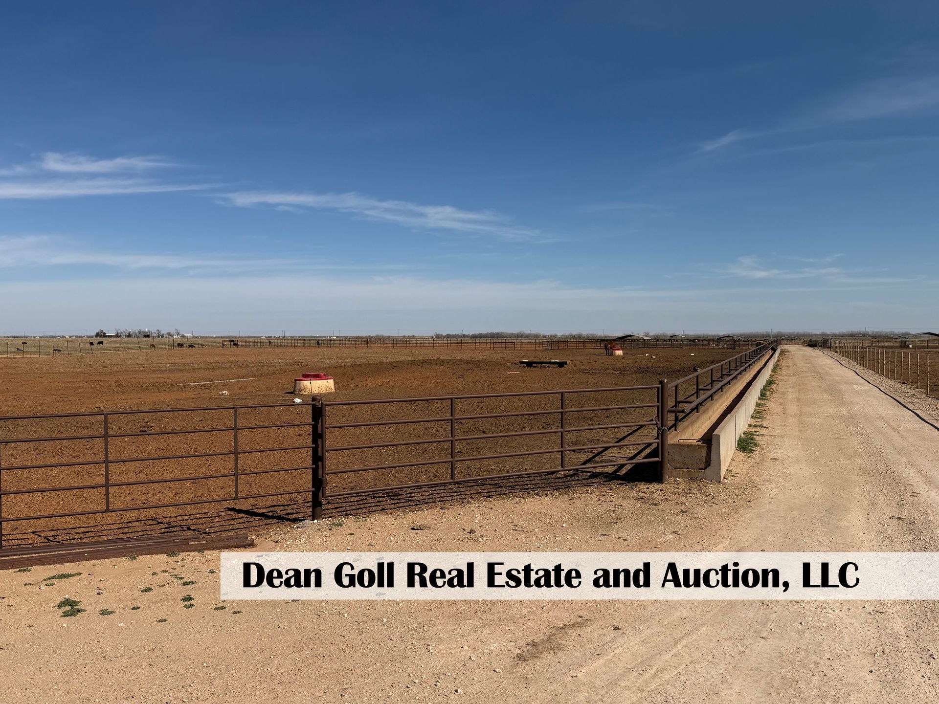 A dirt road runs beside a large, fenced livestock feedlot under a bright blue sky, with a real estate agency watermark.