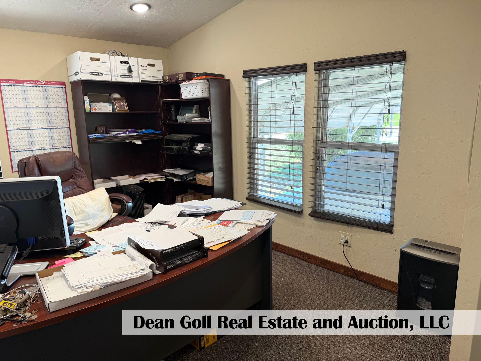 A cluttered office featuring a desk with papers, a monitor, a large bookshelf, two windows with blinds, and a paper shredder.