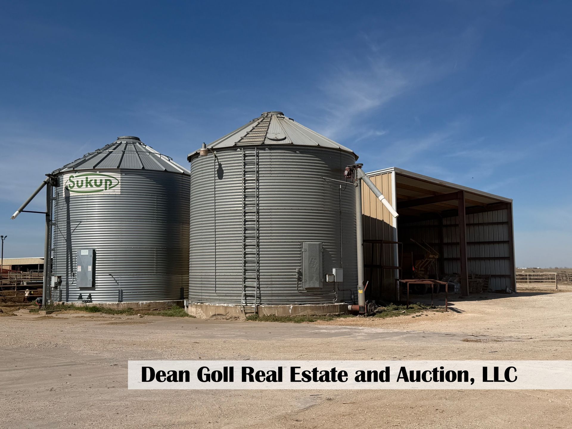 Two grain bins sit next to an open-sided shed under a clear blue sky, with a real estate company name overlaid.