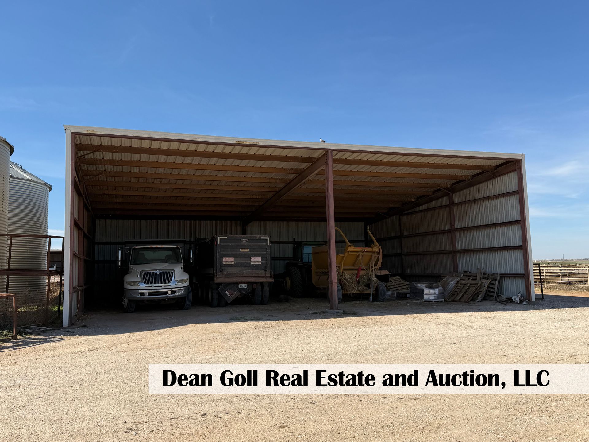A metal storage shed on a gravel lot holding a truck and farm equipment, with a grain bin nearby under a blue sky.