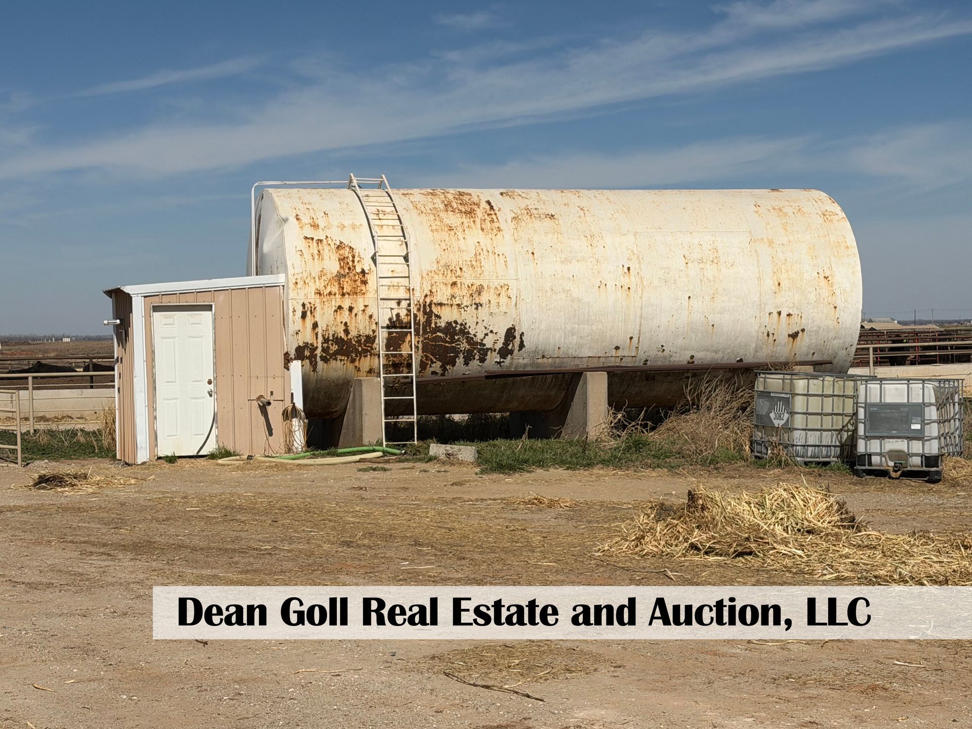 A large, rusted white tank on concrete supports next to a small building, with storage containers on dry, open ground.