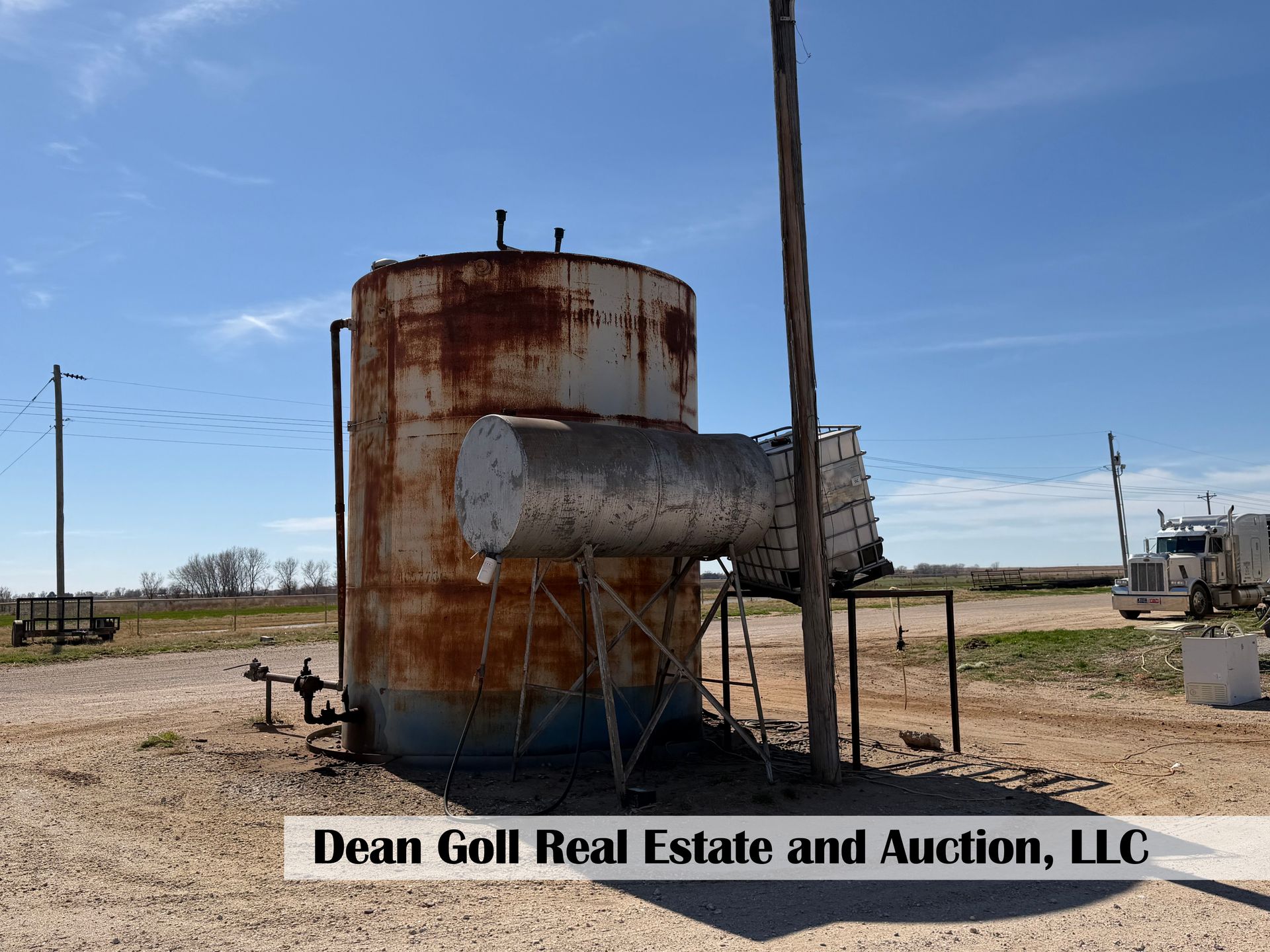 A large, rusted industrial metal storage tank with an attached horizontal tank sitting on a dirt lot under a blue sky.