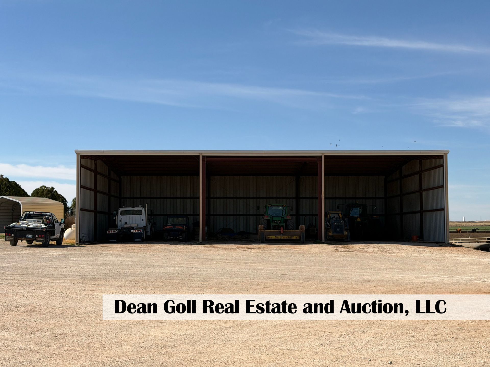 A large, three-bay metal shed stores farm machinery on a gravel lot under a clear blue sky.