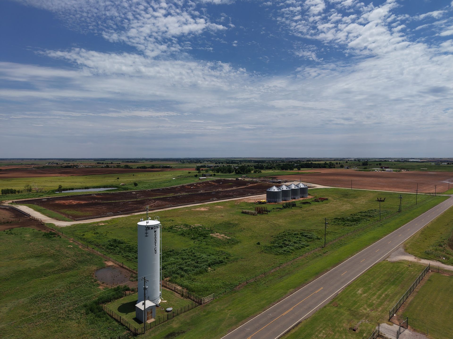 A rural landscape with a white silo, dirt fields, and a road under a partly cloudy sky.