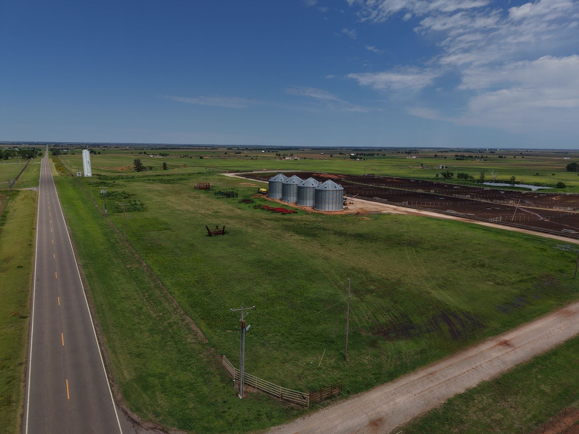 Rural farmland with a road, green fields, grain silos, and a clear blue sky