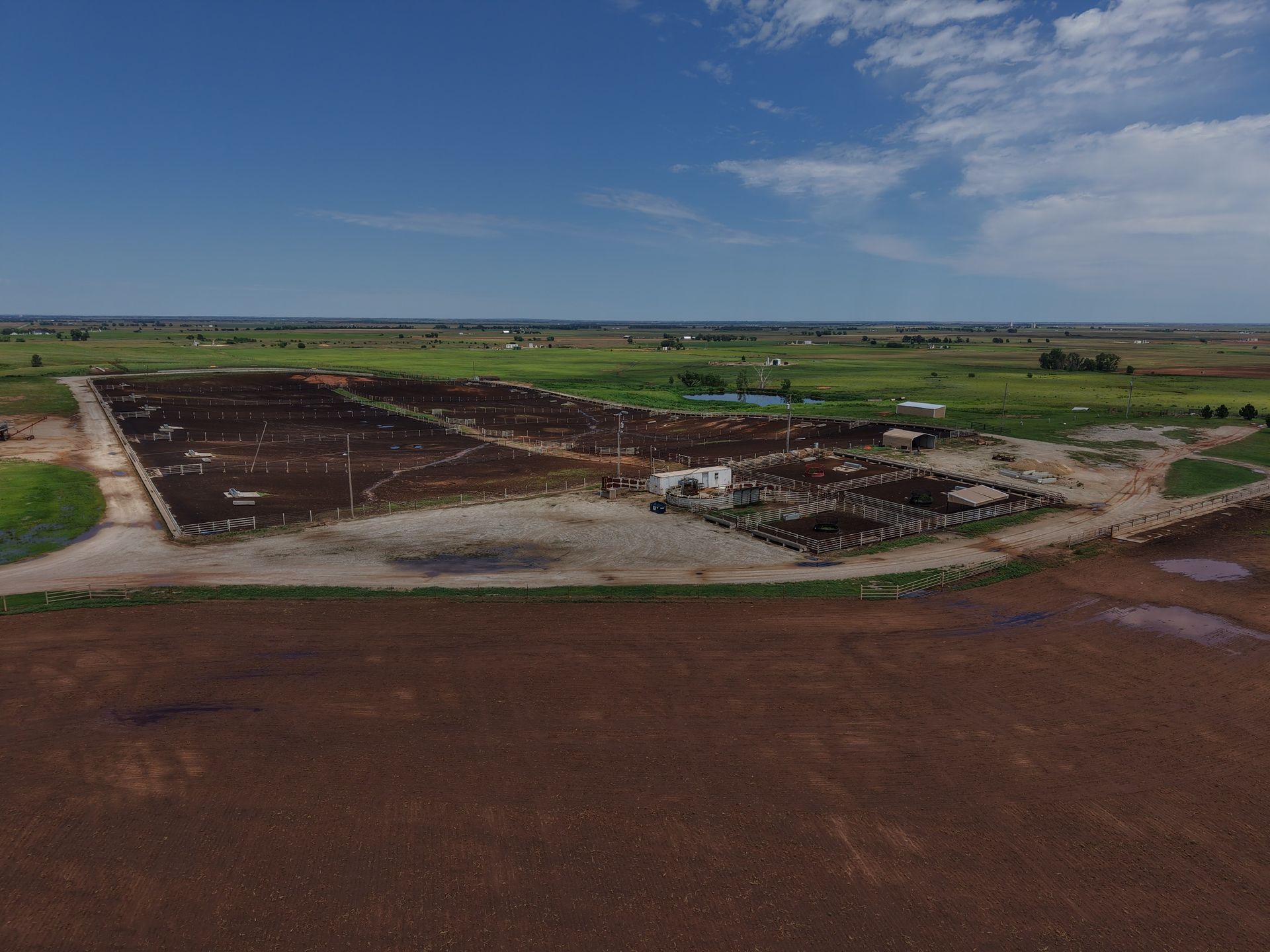 Aerial view of a large farm field with muddy roads, green plots, and a few scattered structures under a blue sky