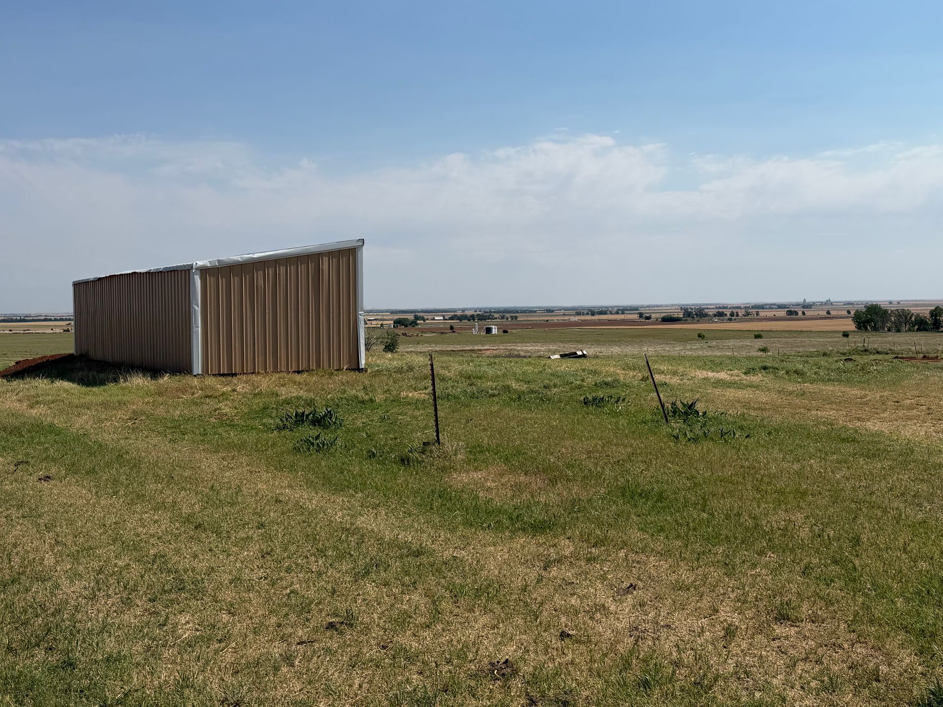 Rural field with a small corrugated metal shed under a blue sky, with distant buildings on the horizon