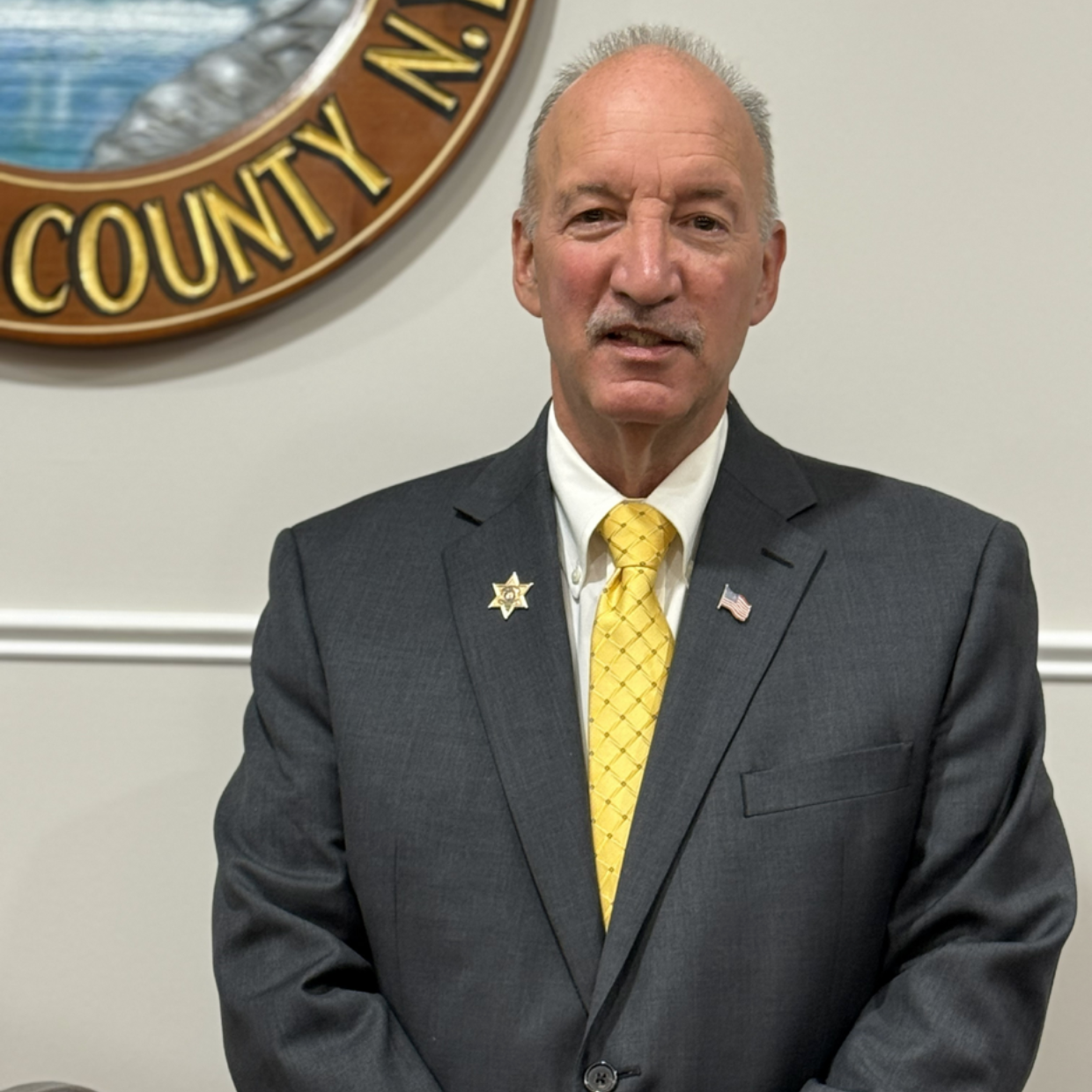 Man in a dark suit and yellow tie standing before a county seal on a white wall.