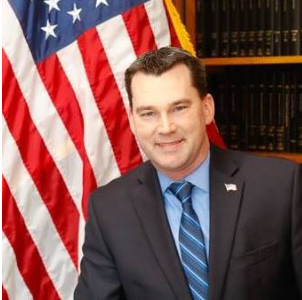 Man in a suit seated before an American flag and bookshelves, smiling at the camera