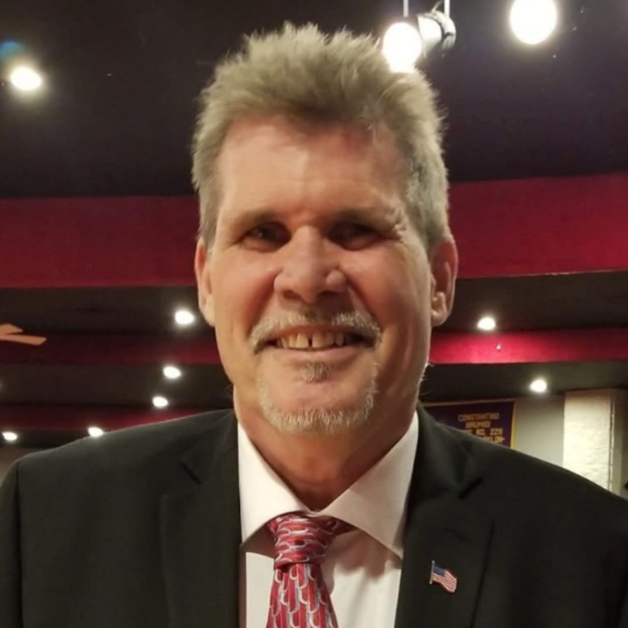Smiling man in a dark suit and red tie at an indoor event with red walls and ceiling lights