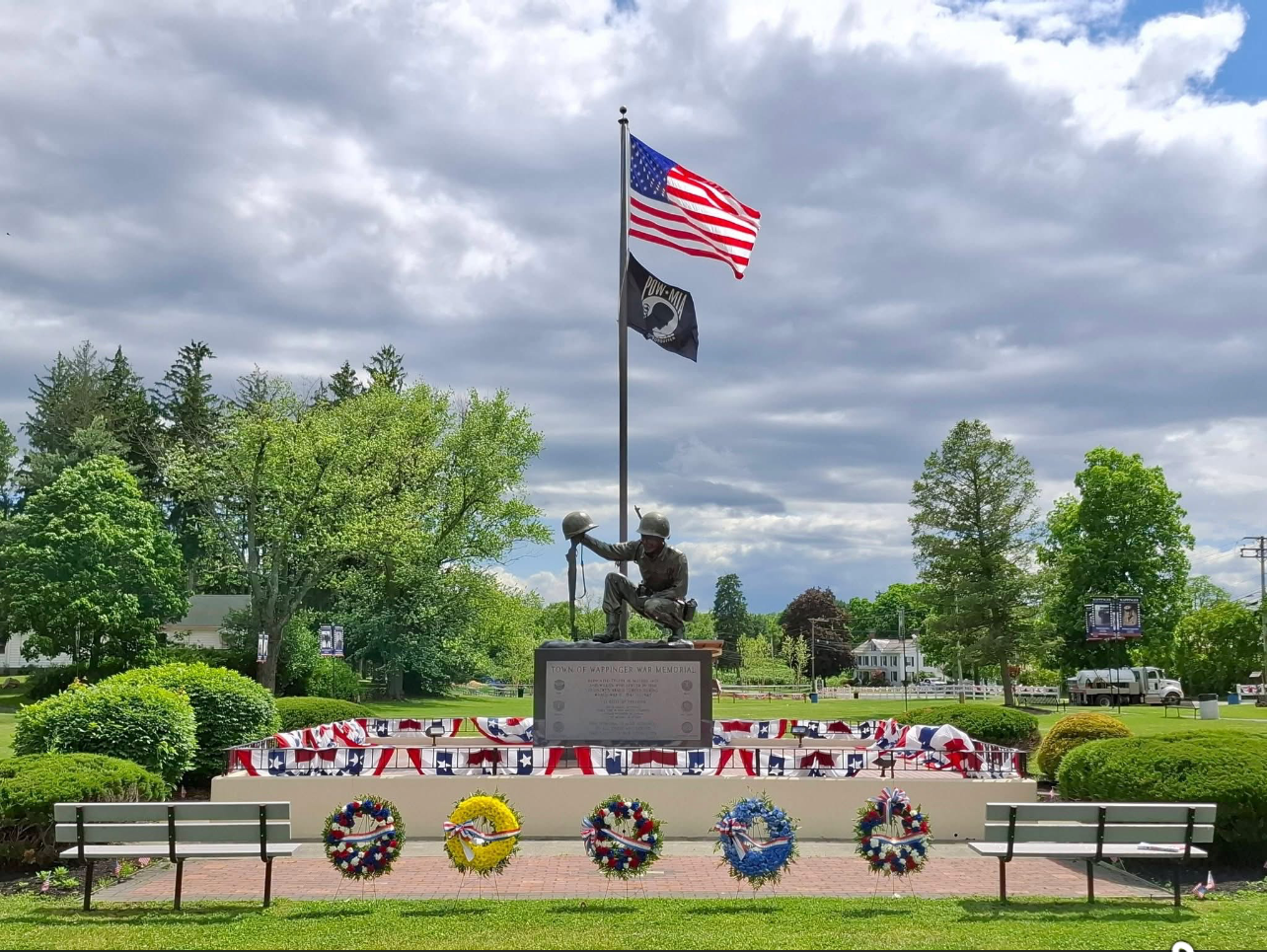 Memorial with soldier statue, U.S. and POW/MIA flags, wreaths, and benches in a park under cloudy sky
