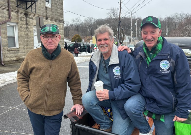 Three men in jackets pose outdoors by a snowy street, one holding a coffee cup.